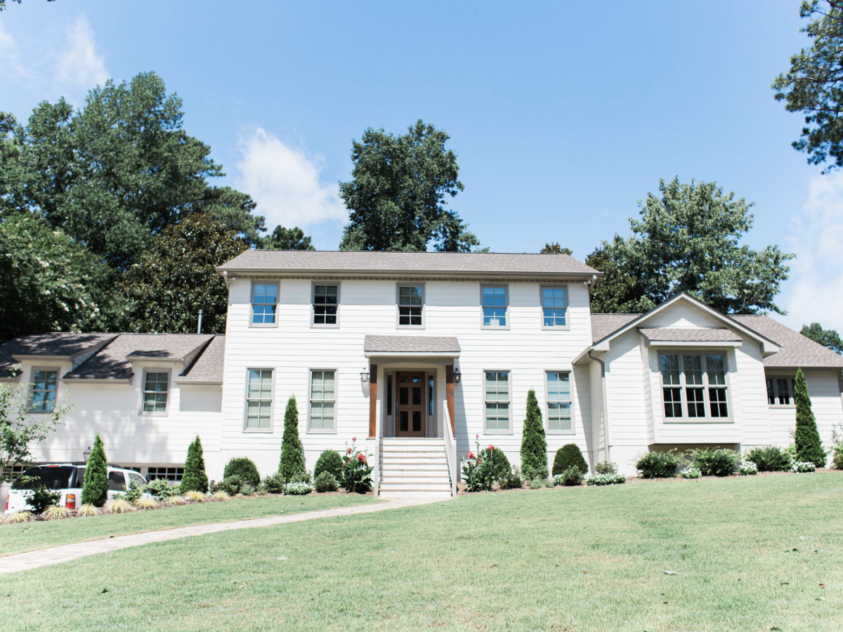 Large two-story white house with a manicured lawn and shrubs, surrounded by tall trees, under a clear blue sky.