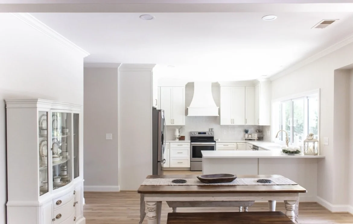 Bright, modern kitchen with white cabinets, stainless steel appliances, a wooden dining table, and a display cabinet.