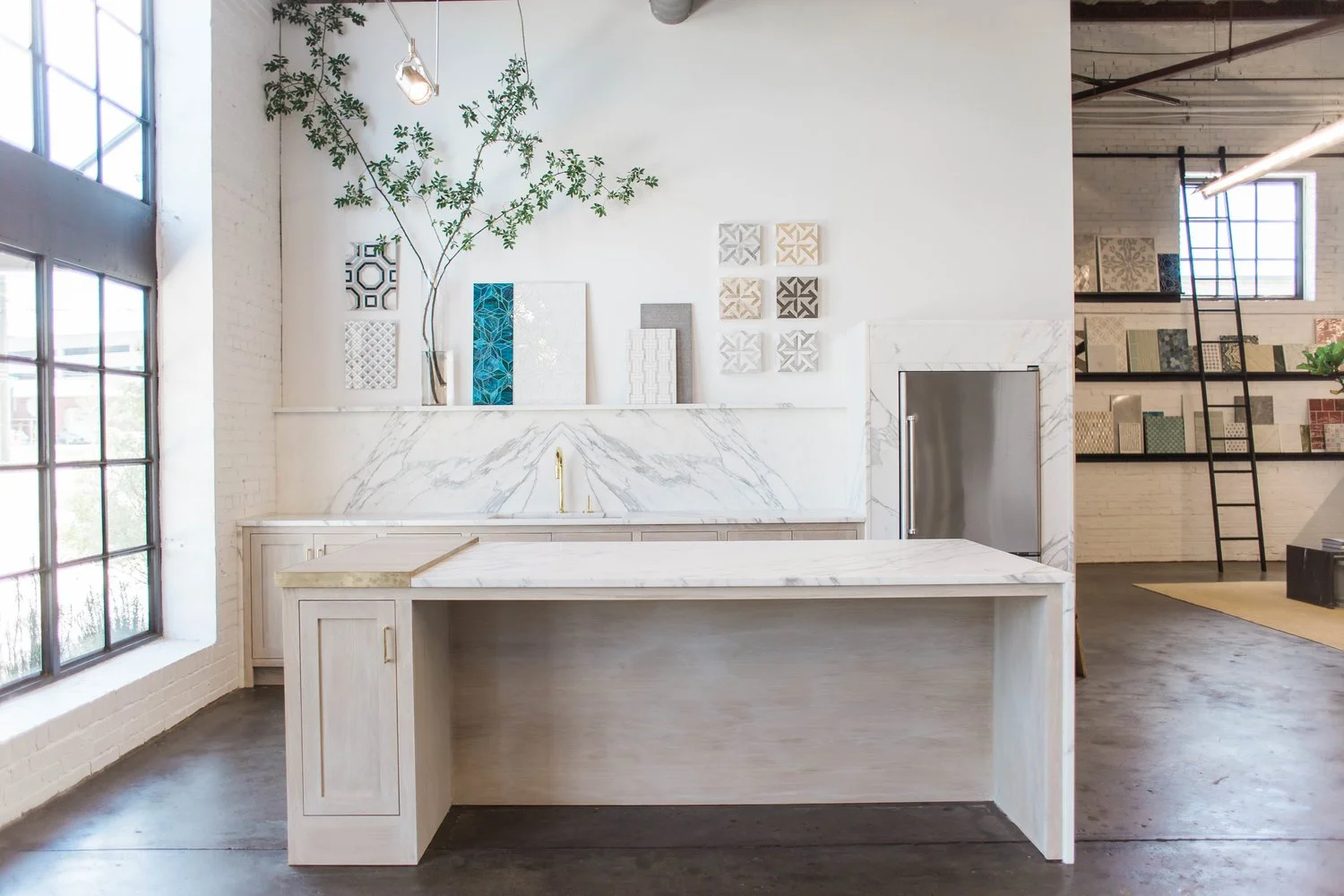 Modern kitchen showroom with white marble island, backsplash, and wall samples, featuring a tree branch decoration and large windows. Downtown Birmingham, AL.