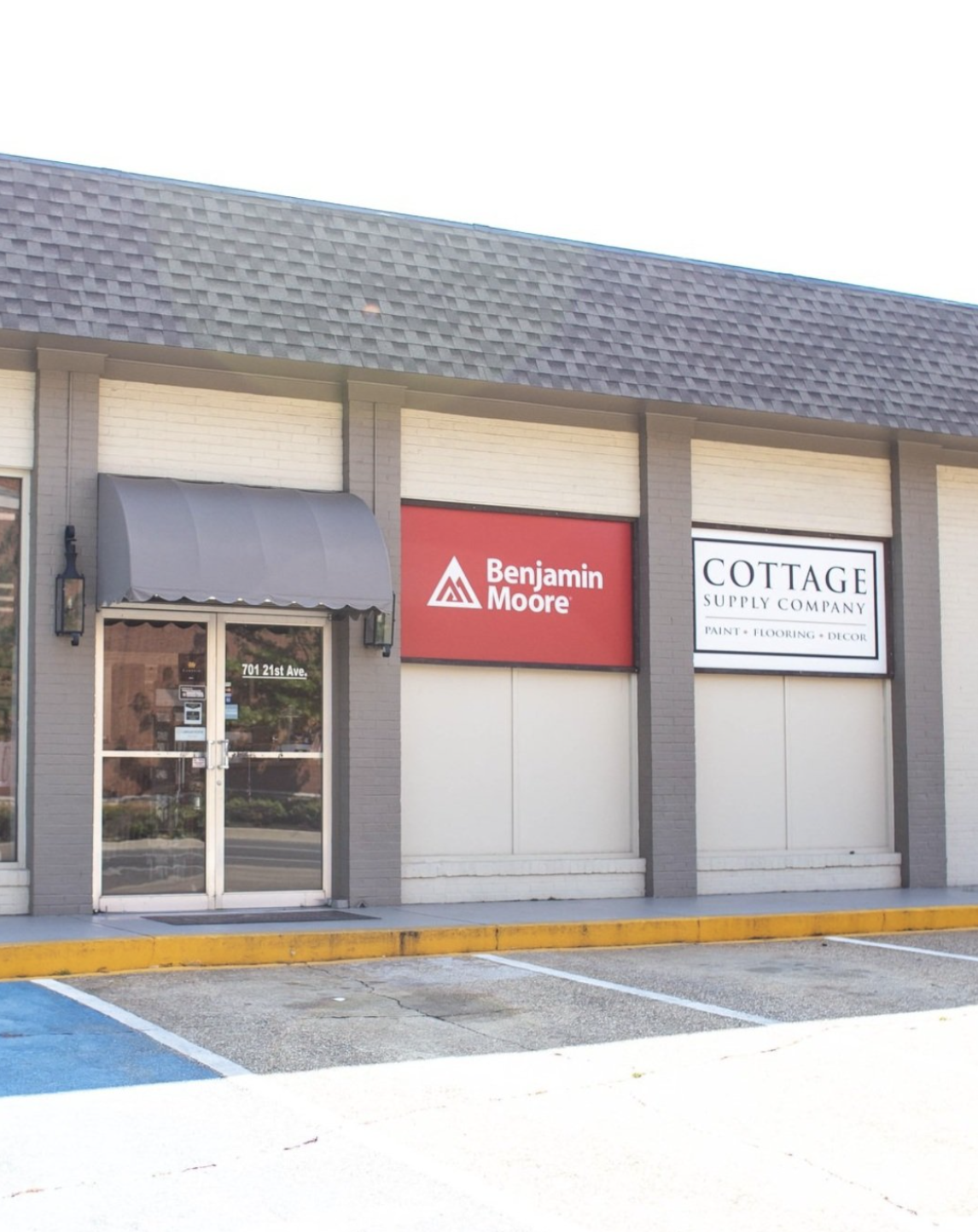 Exterior of a store with Benjamin Moore and Cottage Supply Company signs, featuring a glass door entrance, parking area, and gray awning. Tuscaloosa, AL