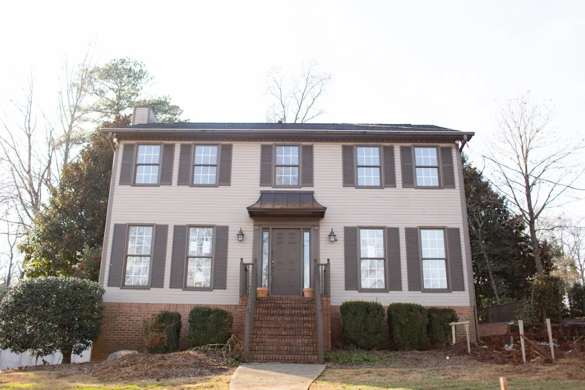 Two-story house with beige siding, brown shutters, and brick foundation, surrounded by small bushes and trees.