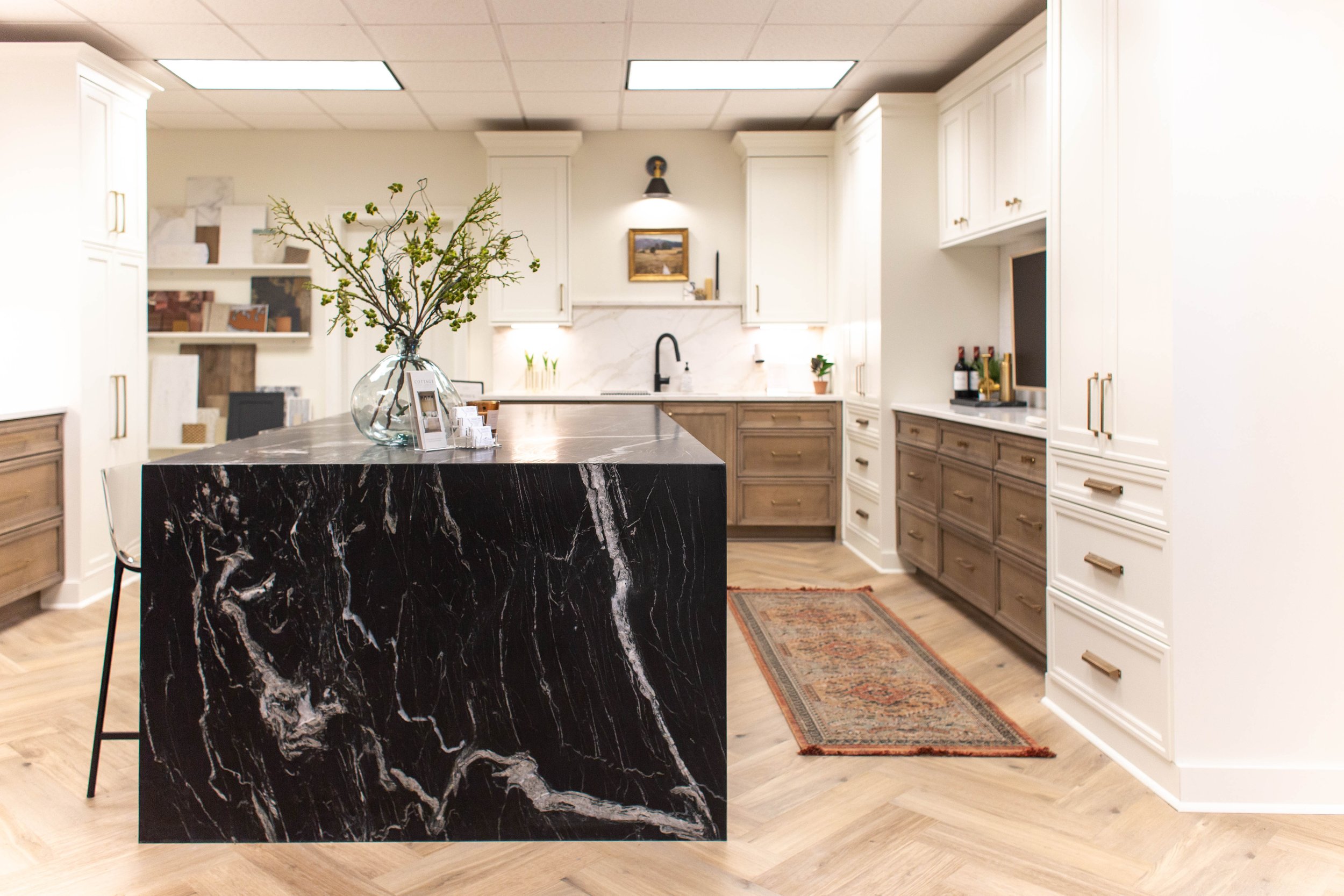Modern kitchen cabinet showroom in Tuscaloosa, AL with white cabinets, black marble island, wooden drawers, decorative rug, potted plants, and vase with branches on countertop.