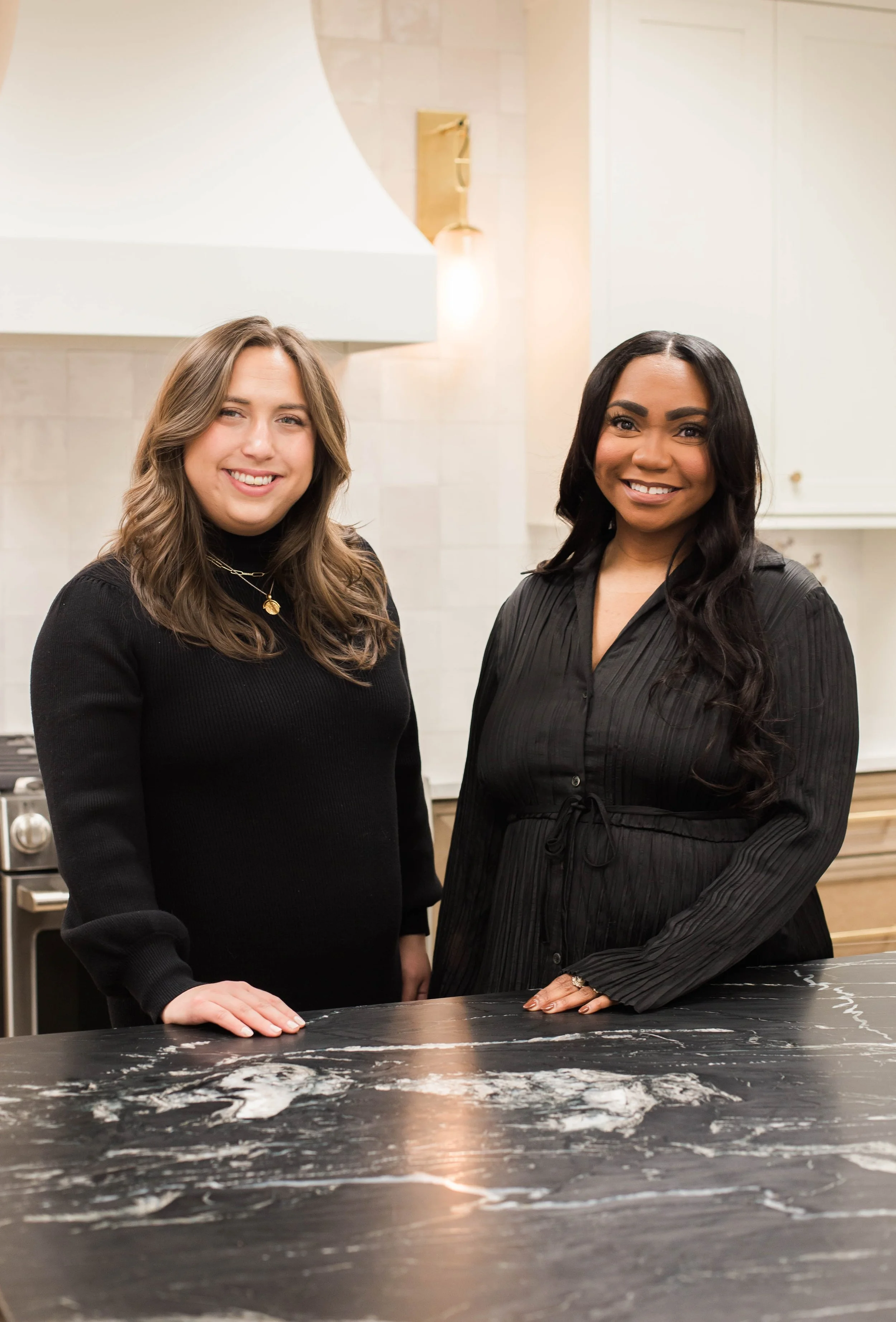 Two women standing behind a black marble countertop in a modern kitchen, both wearing black outfits.