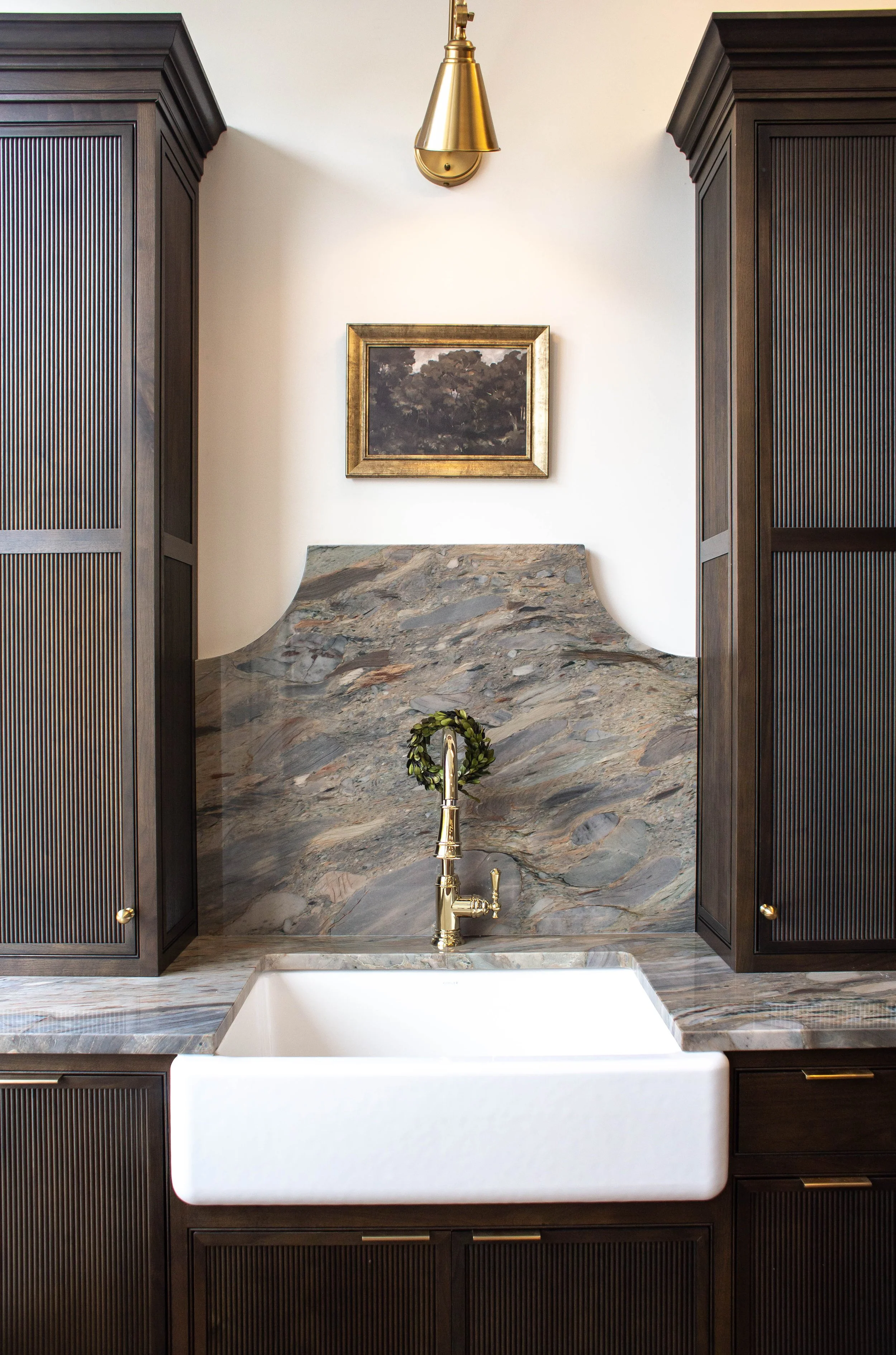Elegant kitchen with marble backsplash, gold faucet, white farmhouse sink, dark wood cabinets, brass sconce, and framed art.