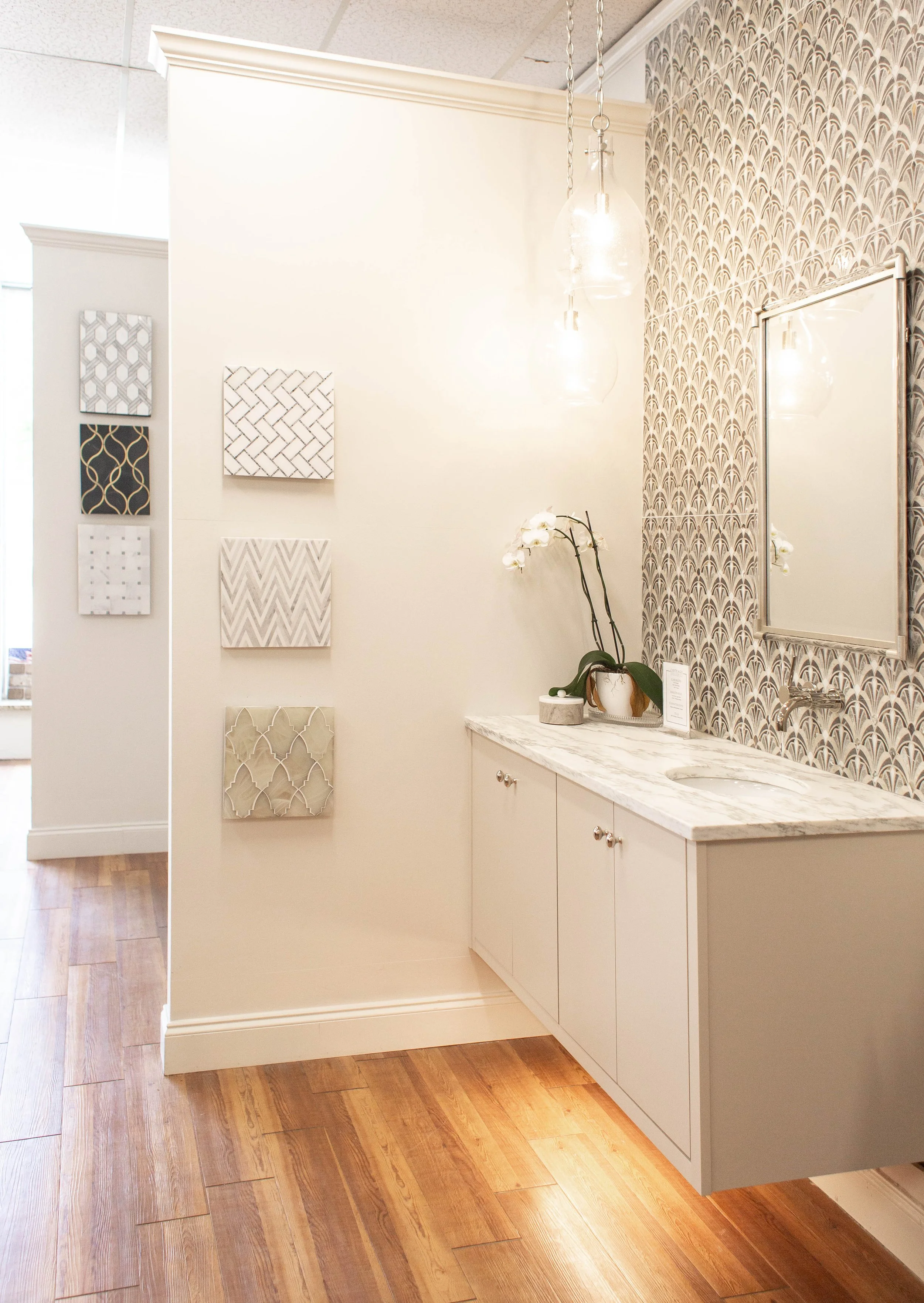 Elegant bathroom interior with modern patterned wallpaper, a marble countertop with a mounted sink, wall-mounted cabinetry, wood flooring, and decorative wall tiles. A potted orchid and pendant lights add to the decor.