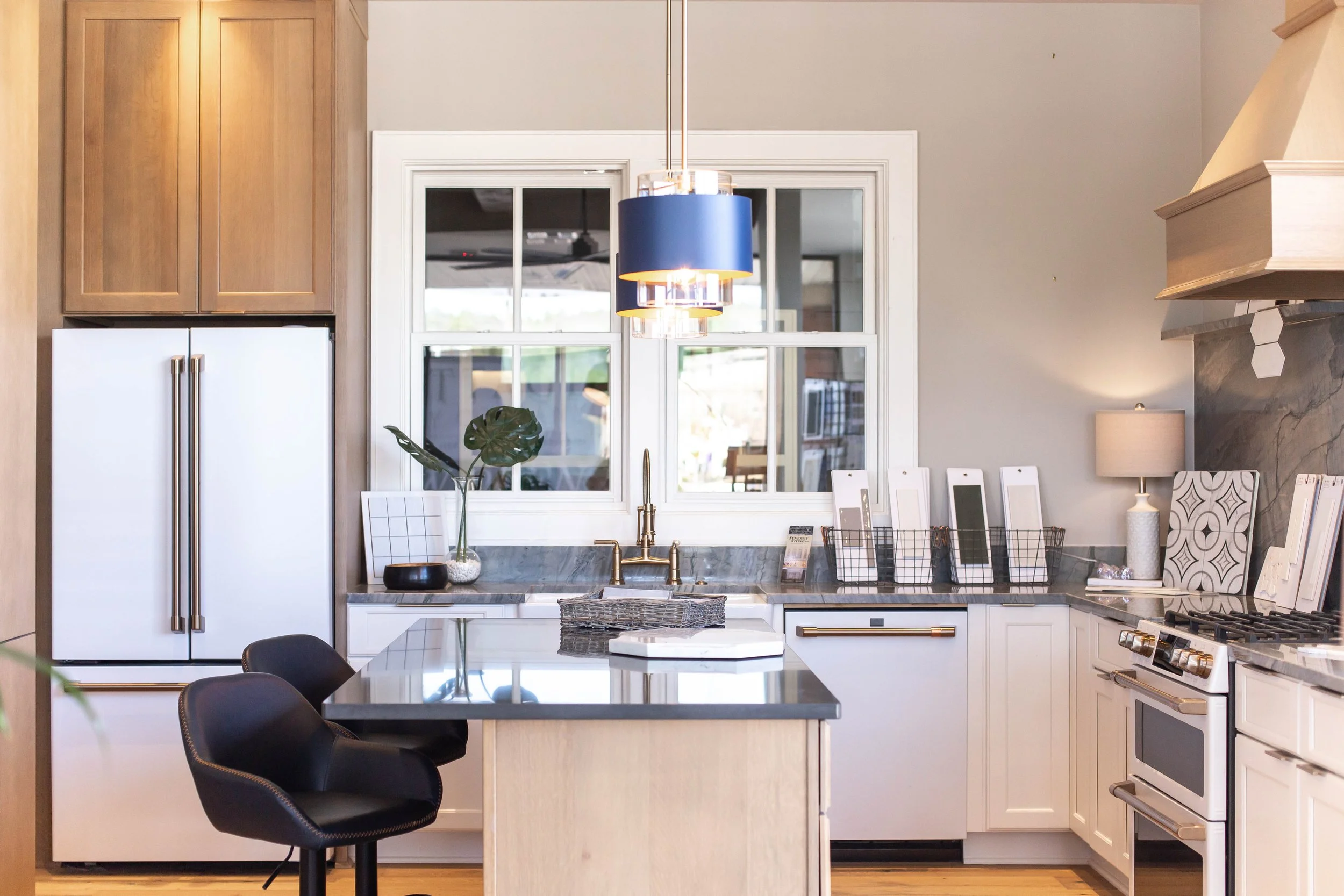 Modern kitchen interior with a central island, black bar stools, white cabinetry, stainless steel appliances, a blue pendant light, and decorative backsplash tiles.