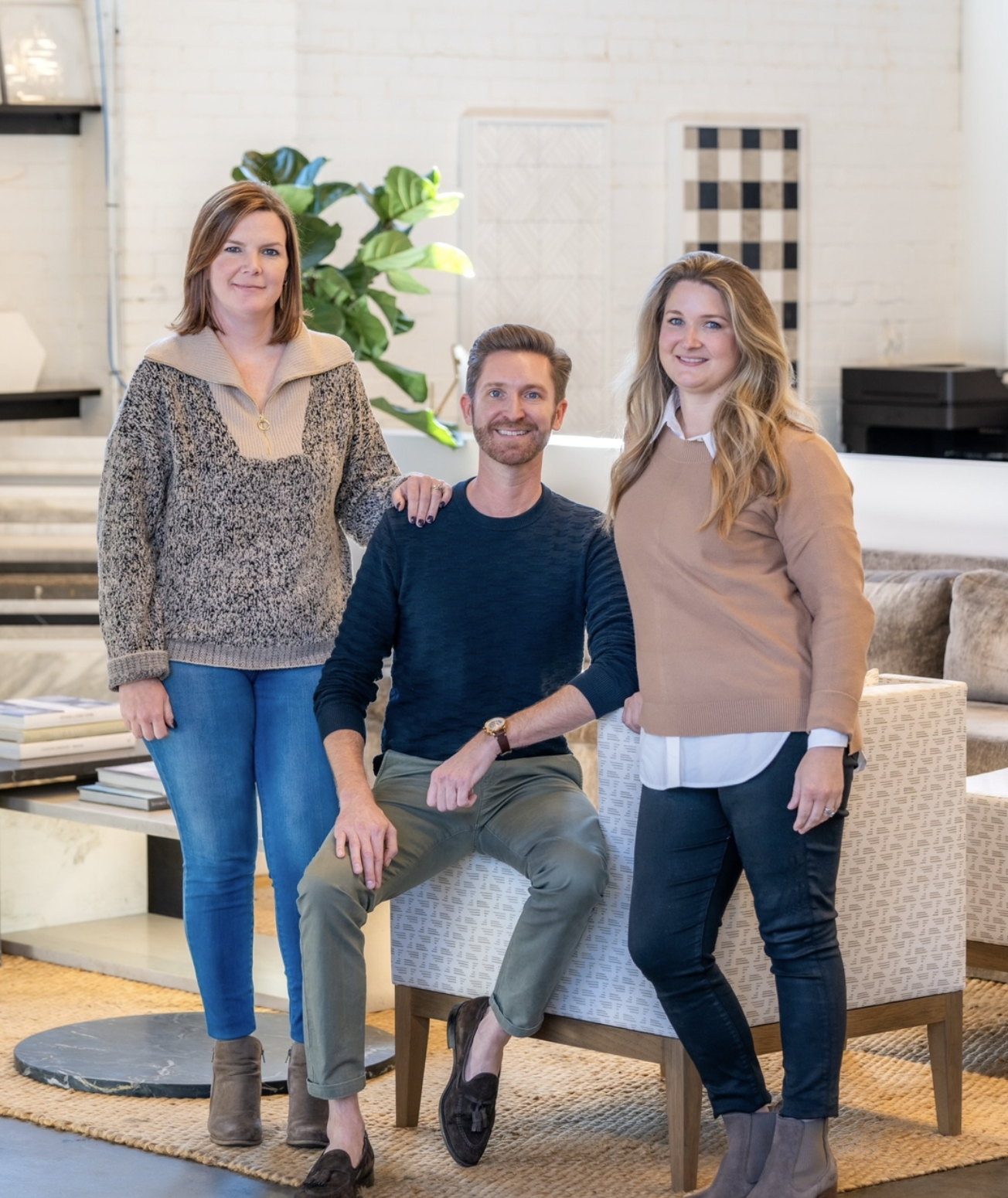 Three people posing in an interior setting with chairs and plants, wearing casual attire.