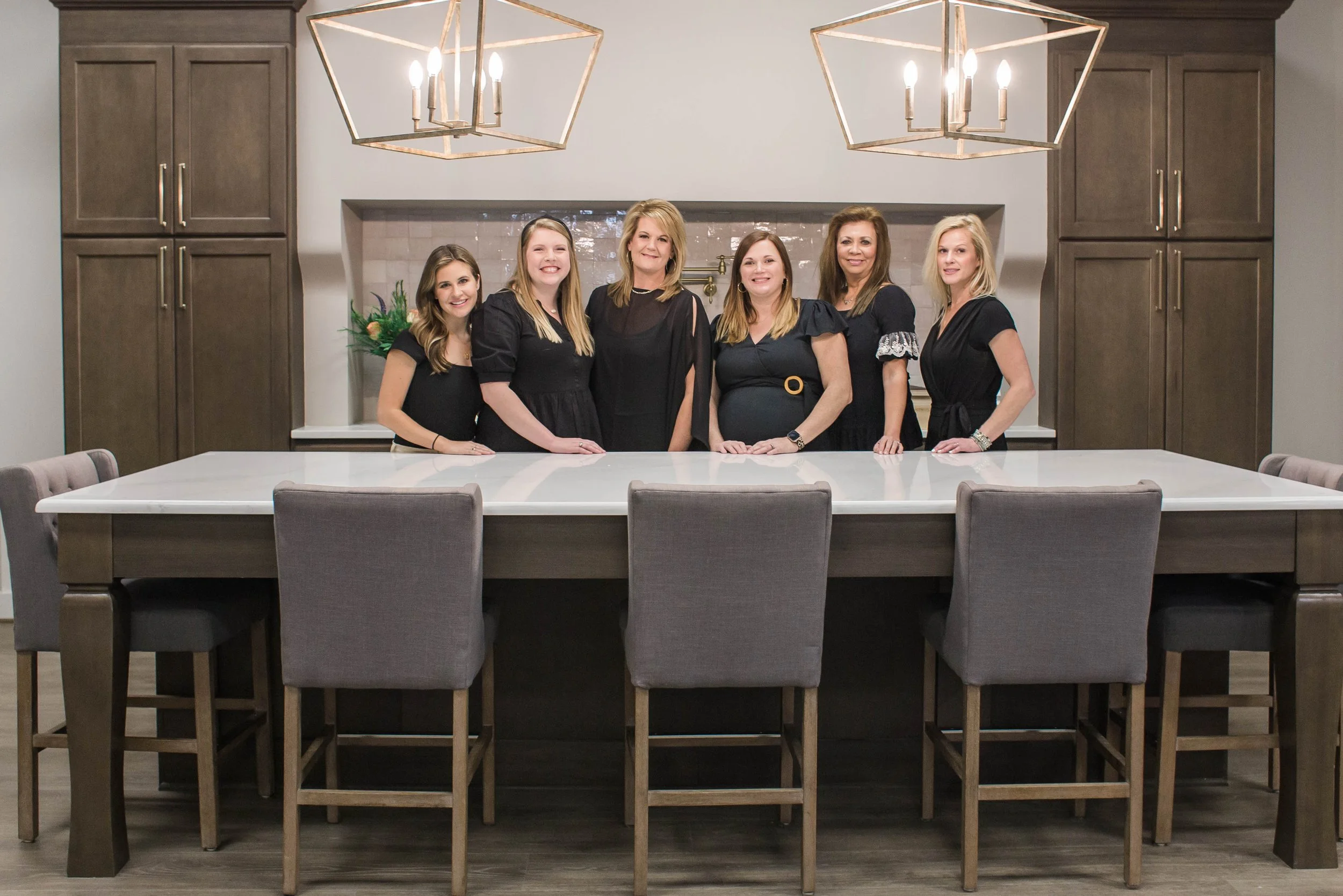 A group of six women standing behind a large kitchen island with chairs, under two geometric pendant lights. Cabinet Showroom in Pelham, AL.
