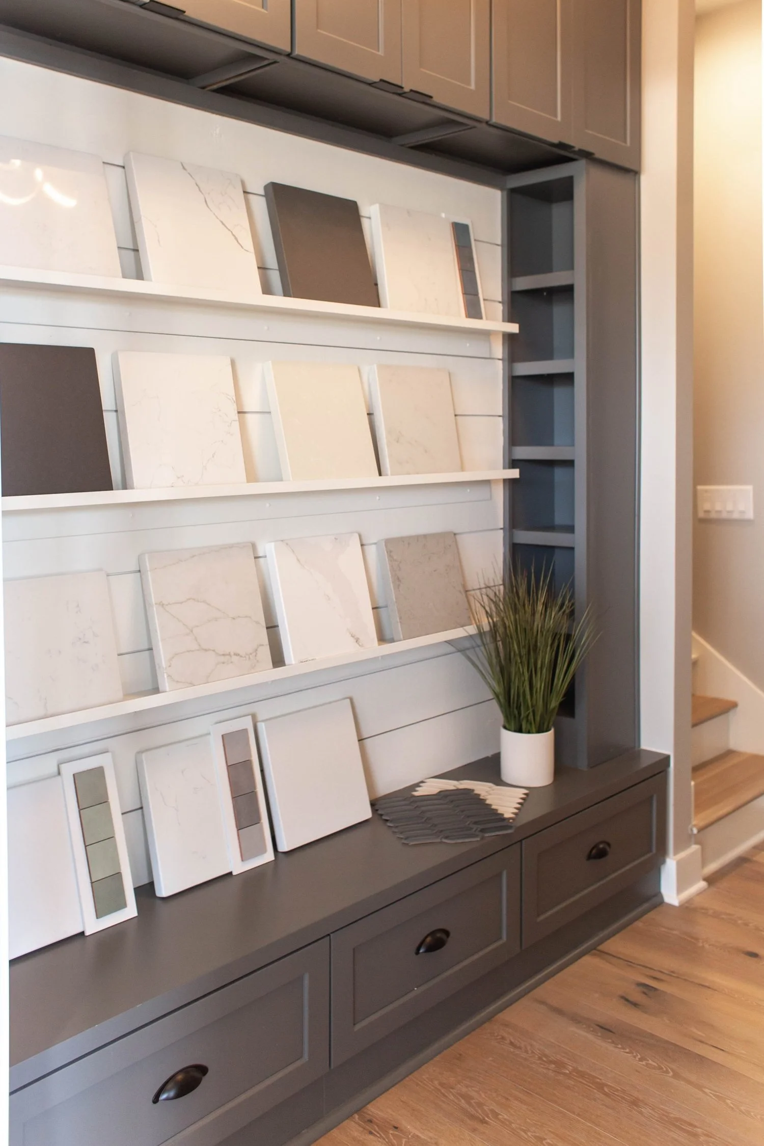 Display of countertop sample panels on shelves in a showroom with grey cabinets and drawers; includes various marble and stone designs, a small plant, and paint swatches on a wooden floor. Cottage Supply Company in Chelsea, AL.