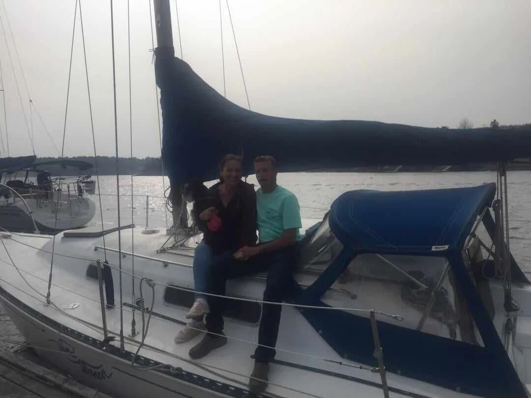 Two people sitting on a sailboat docked near a calm body of water, with another sailboat visible in the background.