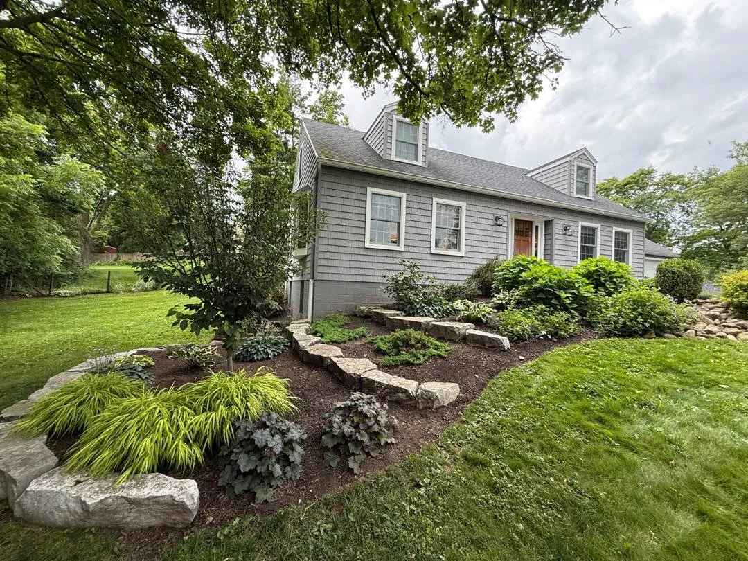 Tiered garden bed with boulder edging, hostas and golden grasses beside a gray home — Genesee Valley Landscape