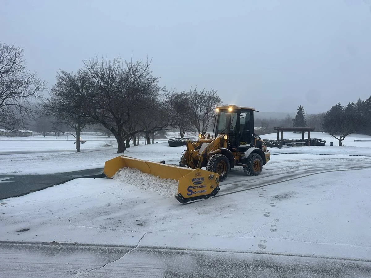 Wheel loader with a sectional snow pusher clearing a commercial lot during snowfall — Genesee Valley Landscape