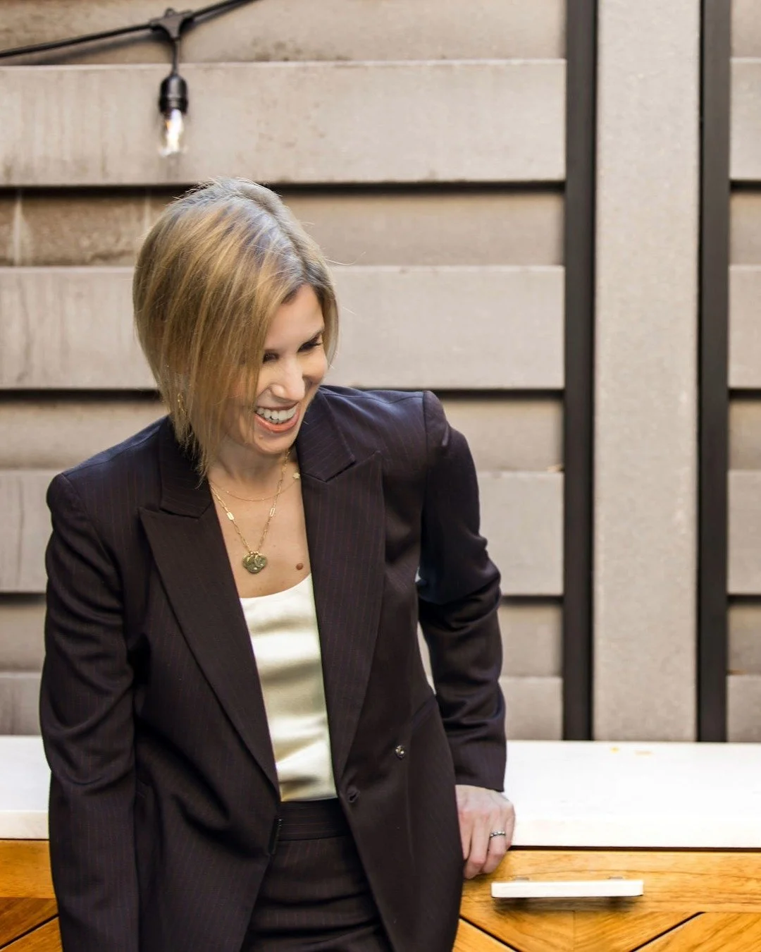 A woman in a dark striped suit and cream blouse, smiling and leaning on a wooden table outdoors.