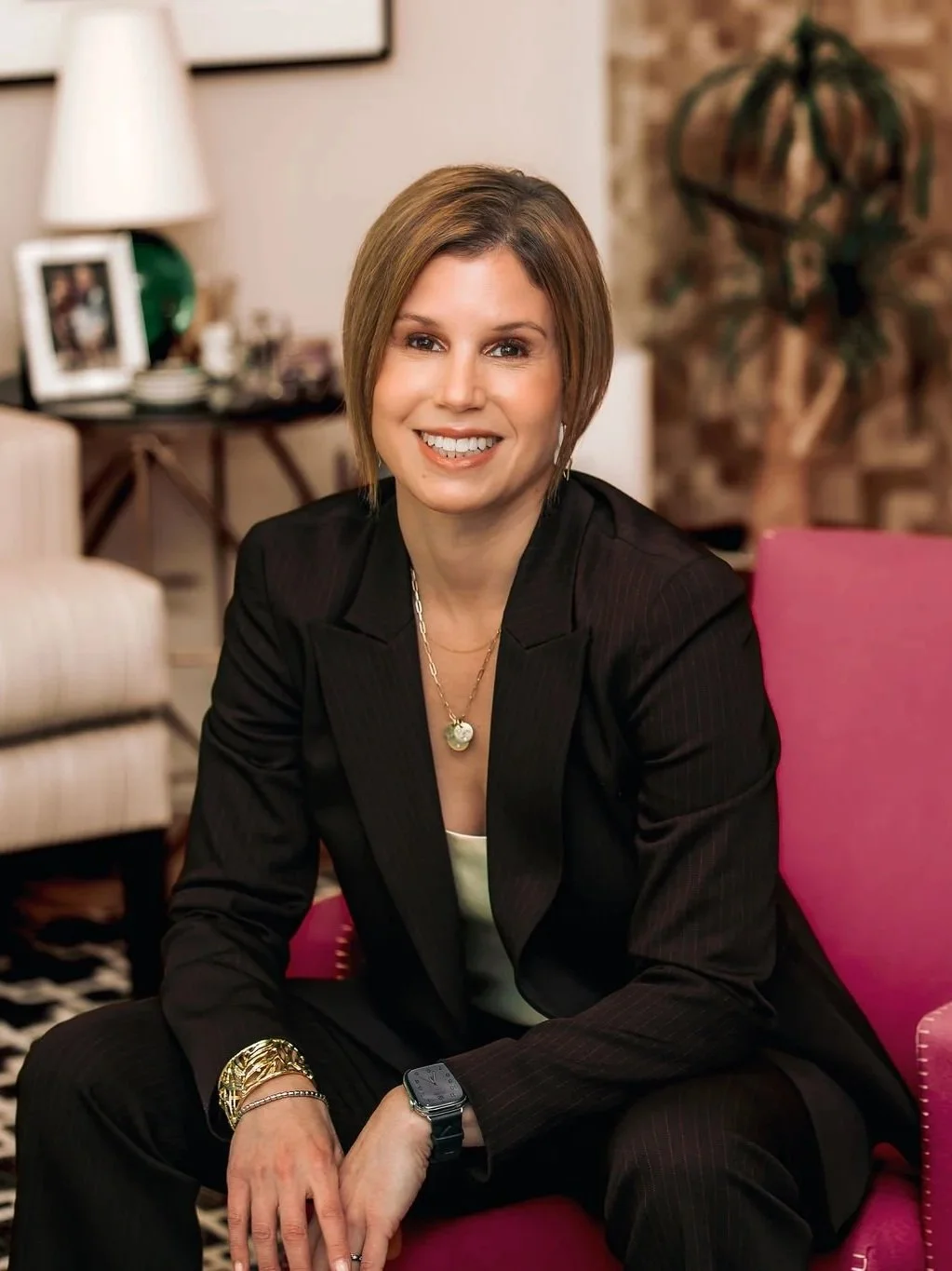 A woman with short brown hair, wearing a black suit and gold jewelry, sitting on a pink chair in a living room with a lamp, picture frames, and a houseplant in the background.