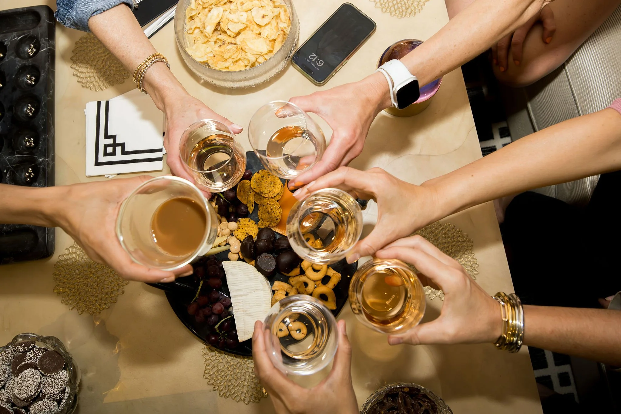 Multiple hands raising glasses of rosé wine for a toast over a table with a cheese platter, crackers, and snacks.