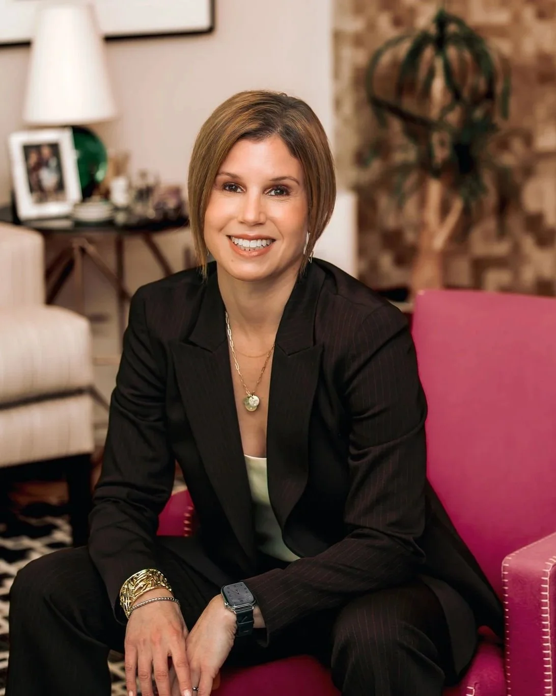 A woman with short brown hair smiling, sitting on a pink chair in a cozy room with a brick wall, a potted plant, and a table with framed photos and a lamp in the background.