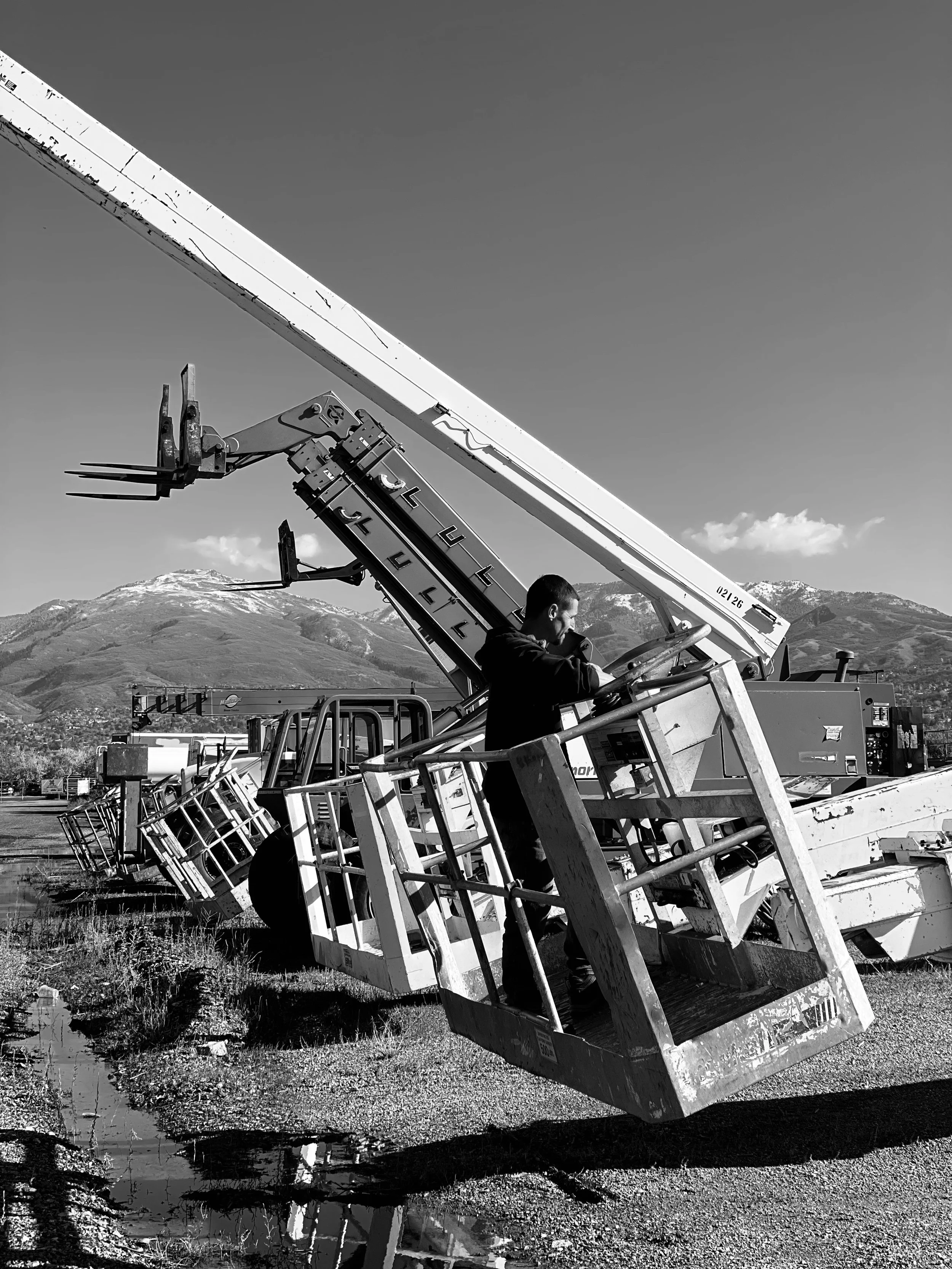 Black and white photo of several mobile cranes stacked outdoors, with mountains in the background and a puddle on the ground reflecting the cranes.