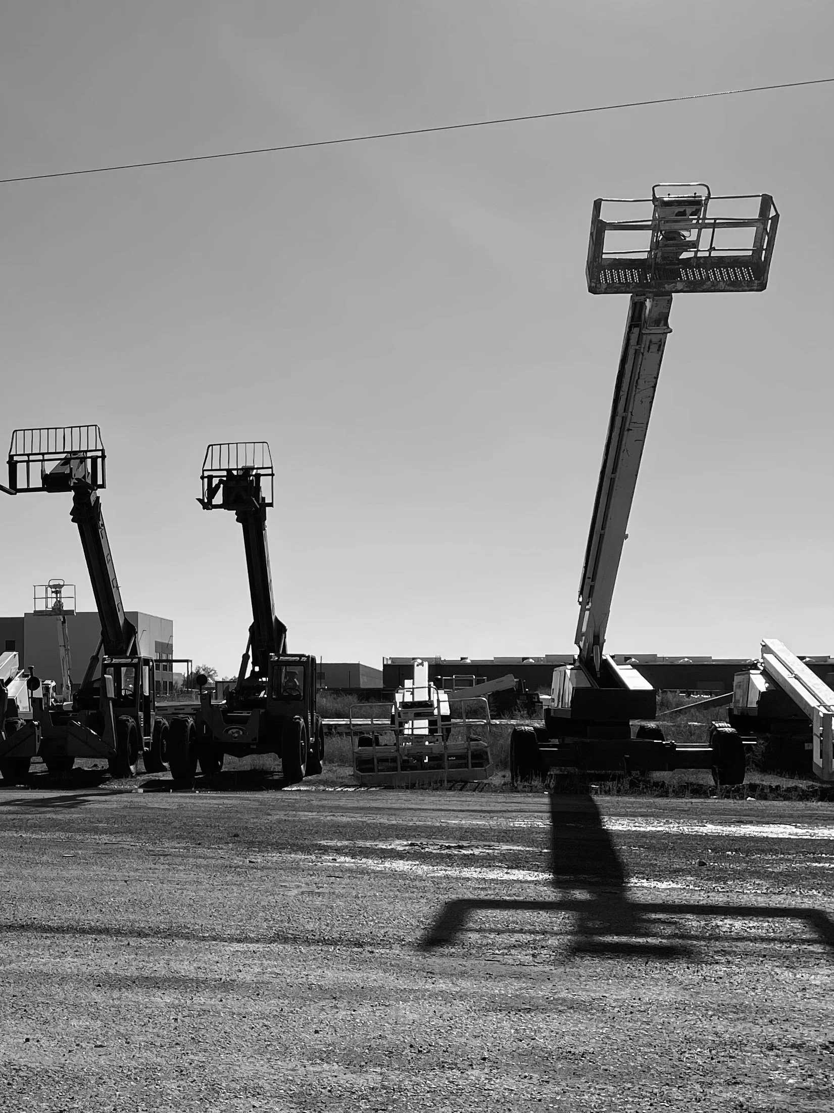 Black and white photo of construction lifts with extended arms, positioned on a gravel lot, with a building and trees in the background and a wire crossing the sky.