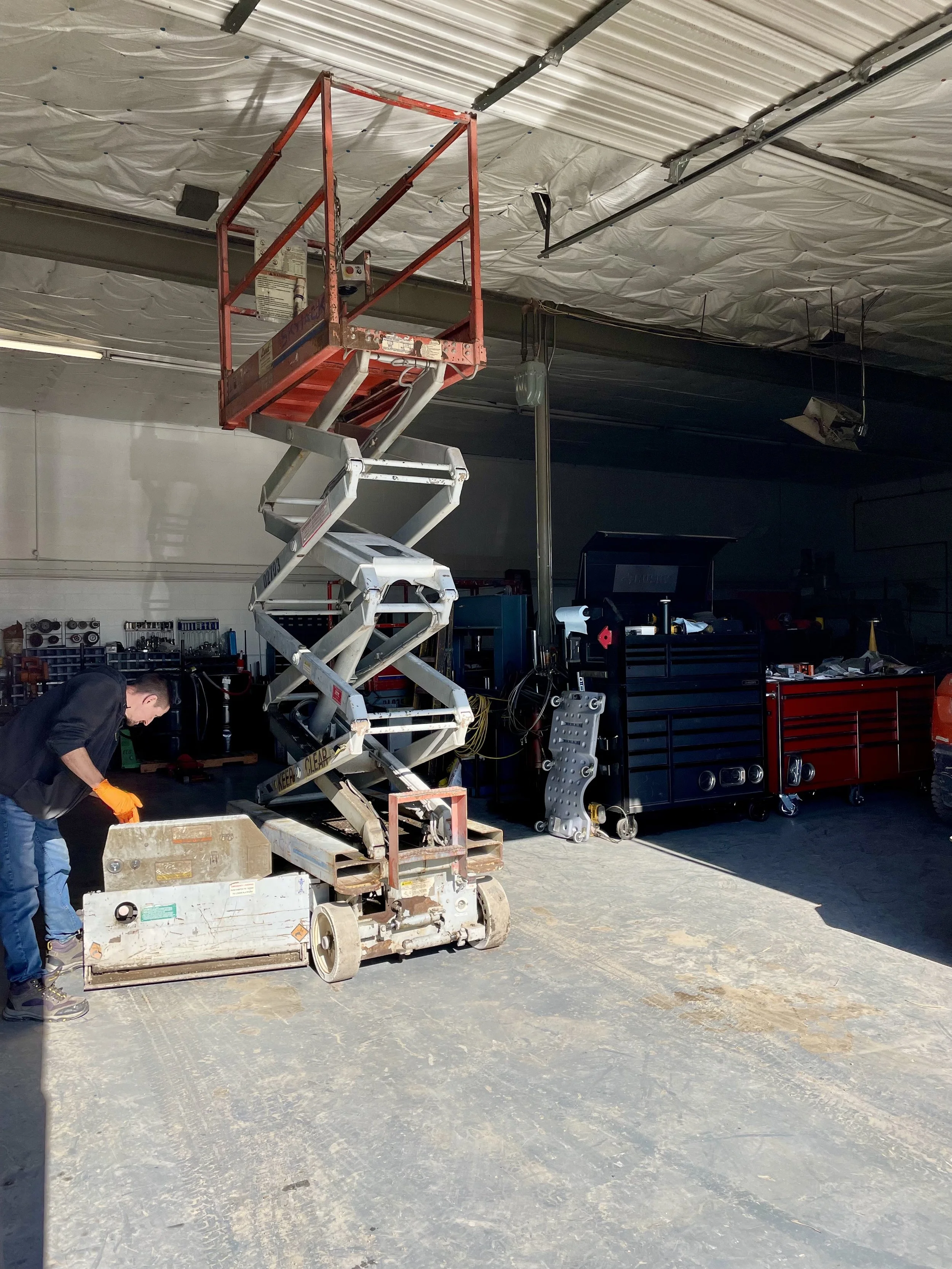 A worker operating a scissor lift inside a warehouse or garage. The lift is raised, and the worker is wearing safety gloves and gloves. The ceiling has insulation material, and there is a tool chest, equipment, and tools organized against the wall.