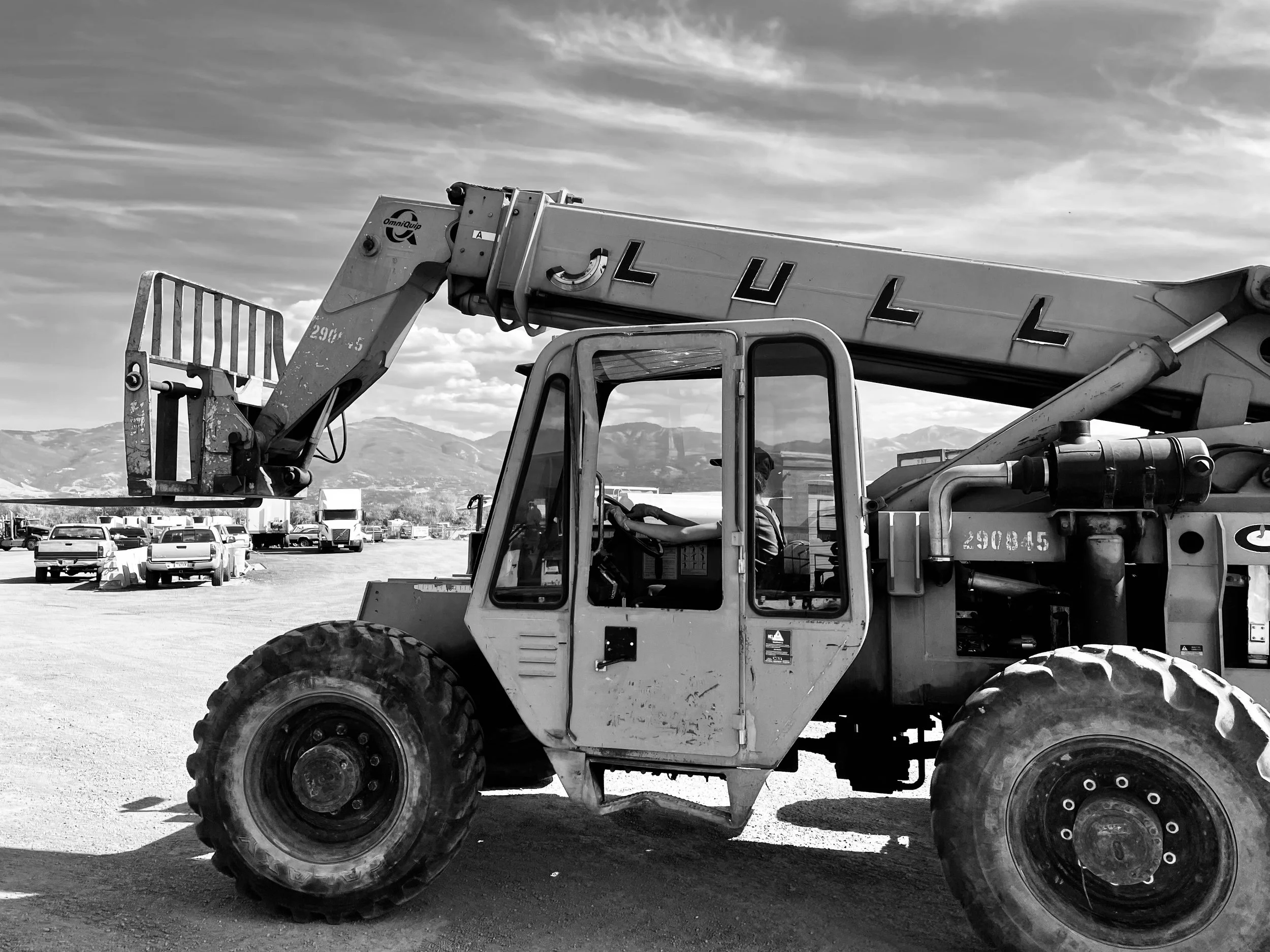 A construction vehicle, specifically a telescopic boom lift, parked on a gravel lot with a backdrop of mountains and a cloudy sky. The vehicle has large tires and a cabin with glass windows.