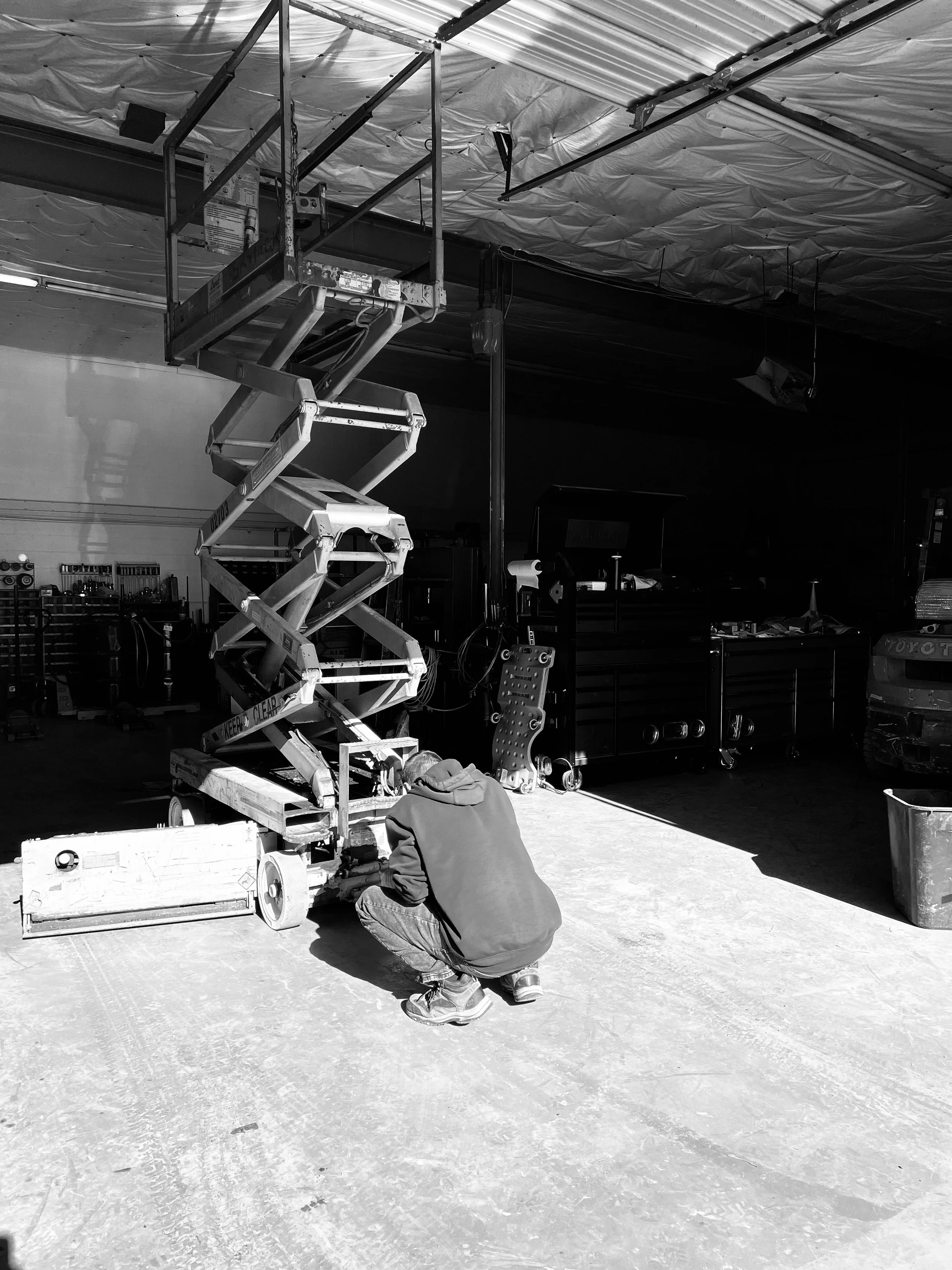 A person crouching in a workshop, working on a mechanical lift or lift platform with a scissor mechanism, in a large industrial space with tools and equipment in the background.