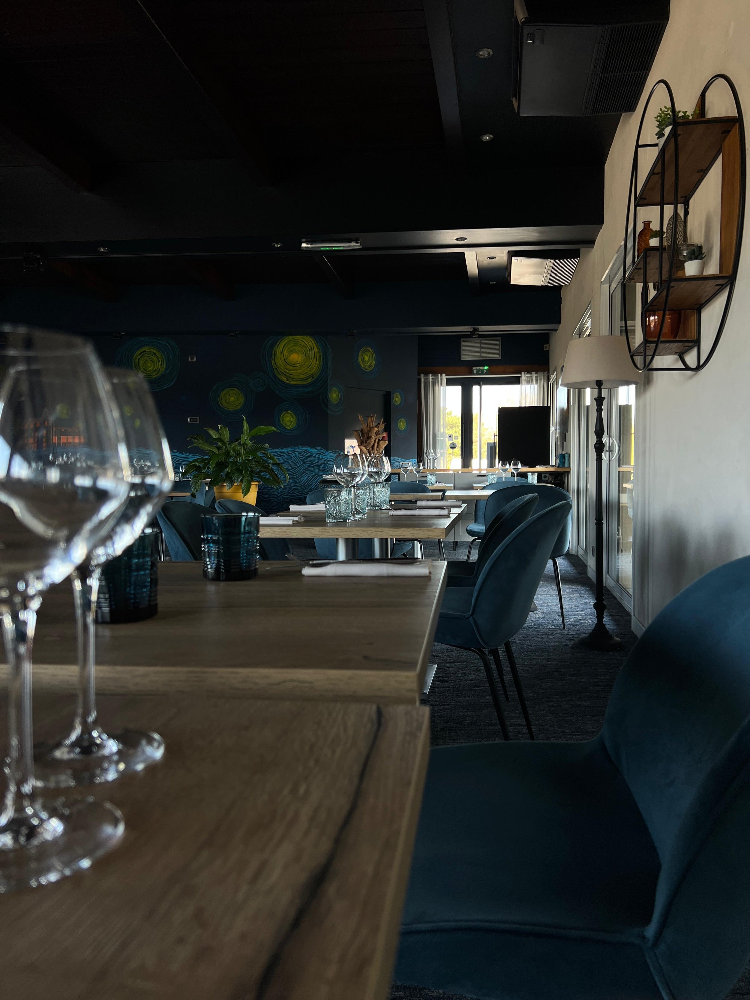 Intérieur d'un restaurant avec des chaises bleues, des tables en bois, des verres à pied, une étagère murale décorative et une peinture colorée sur le mur.