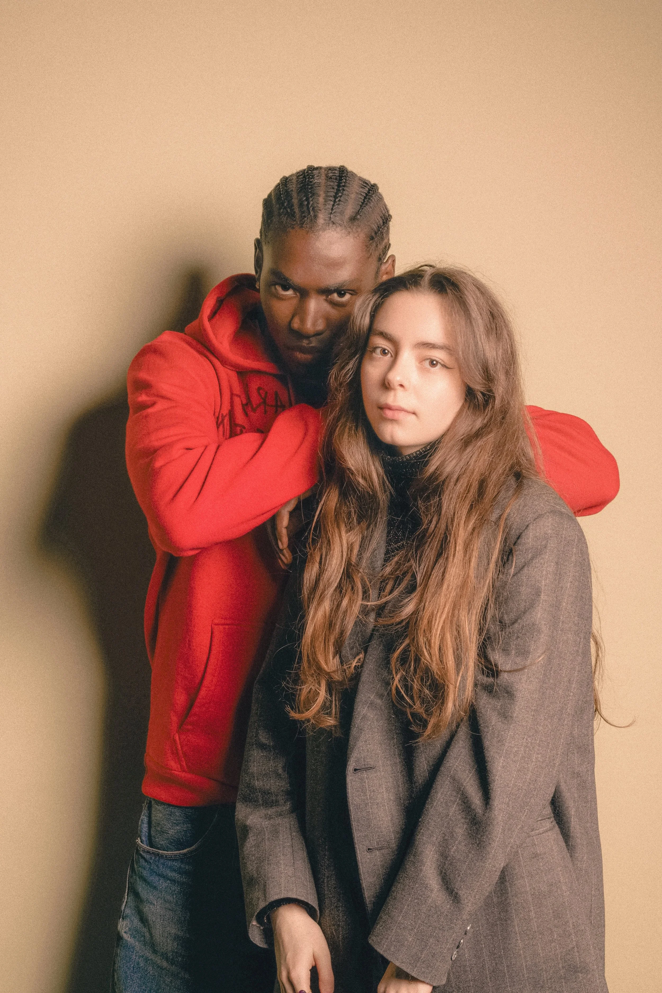 Un homme portant une veste rouge entoure ses bras autour d'une femme vêtue d'un manteau sombre, posant devant un fond neutre.