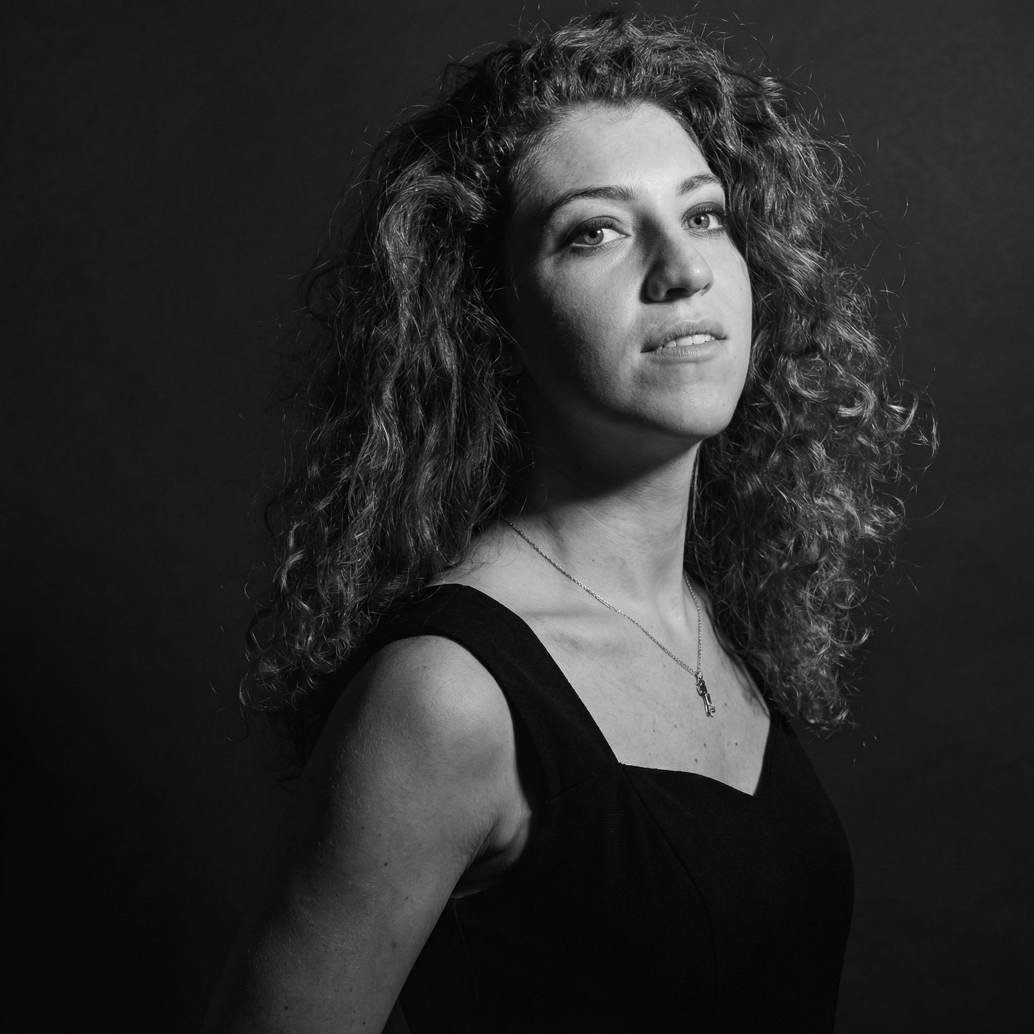 Portrait en noir et blanc d'une femme avec cheveux bouclés, portant un haut noir et une chaîne avec une petite croix