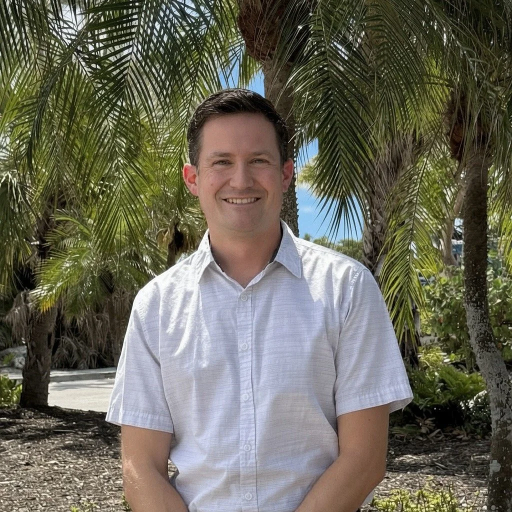 Smiling man with palm trees behind him.