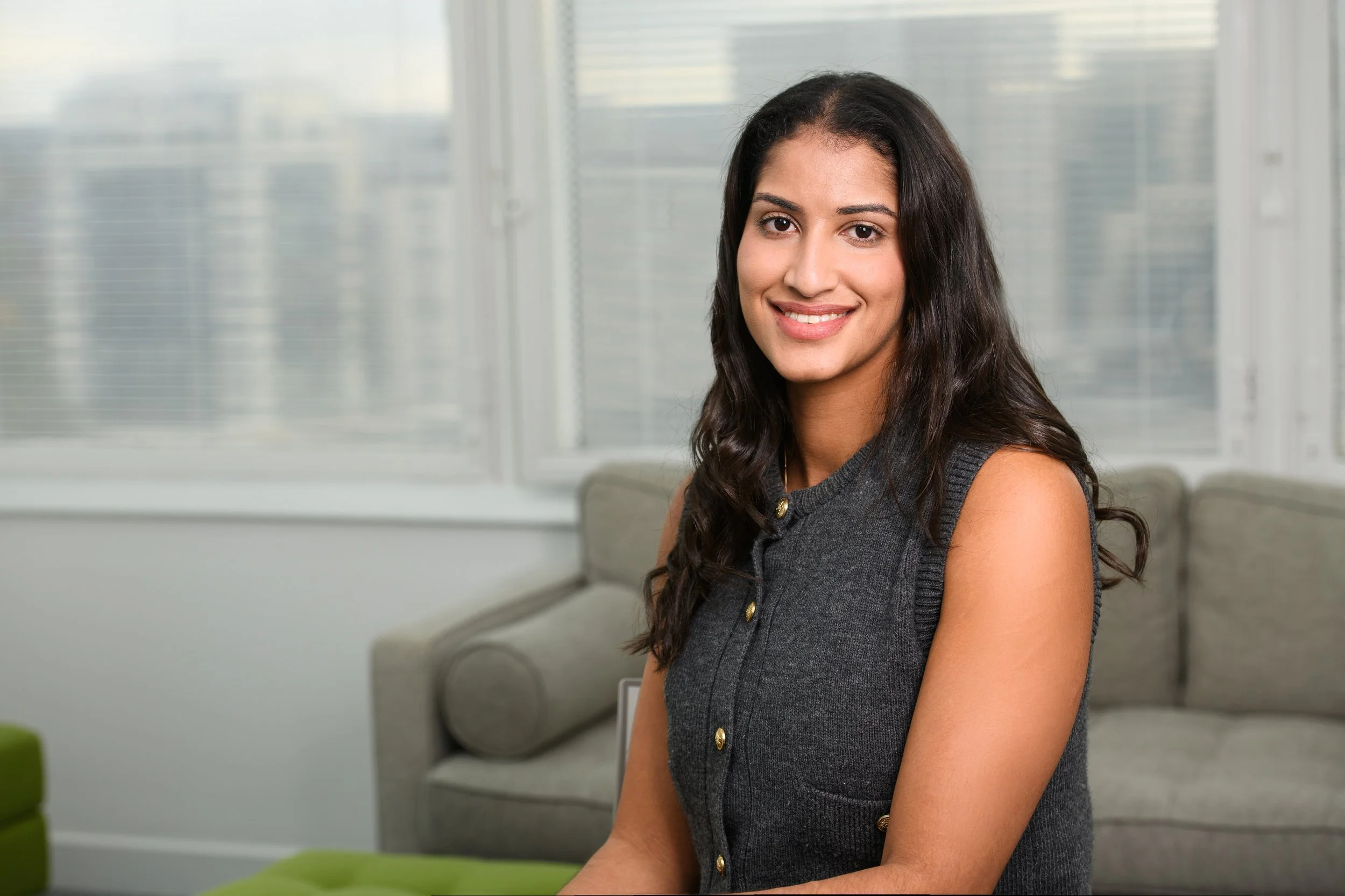 A young woman with long dark hair, wearing a sleeveless dark gray top with gold buttons, smiling in an office setting with large windows and a gray sofa in the background.