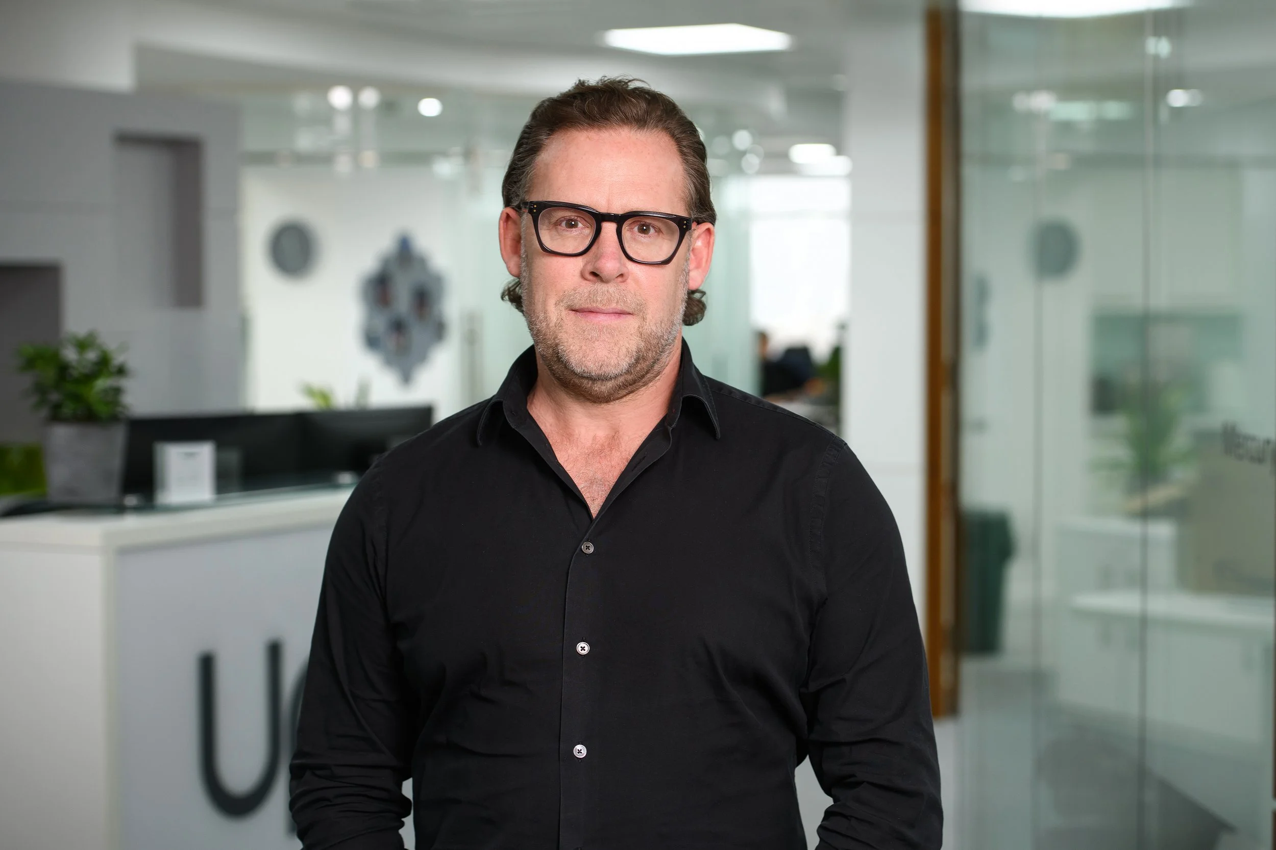A middle-aged man with dark hair, glasses, and light stubble, wearing a black shirt, standing in a modern office environment with glass walls and plants in the background.