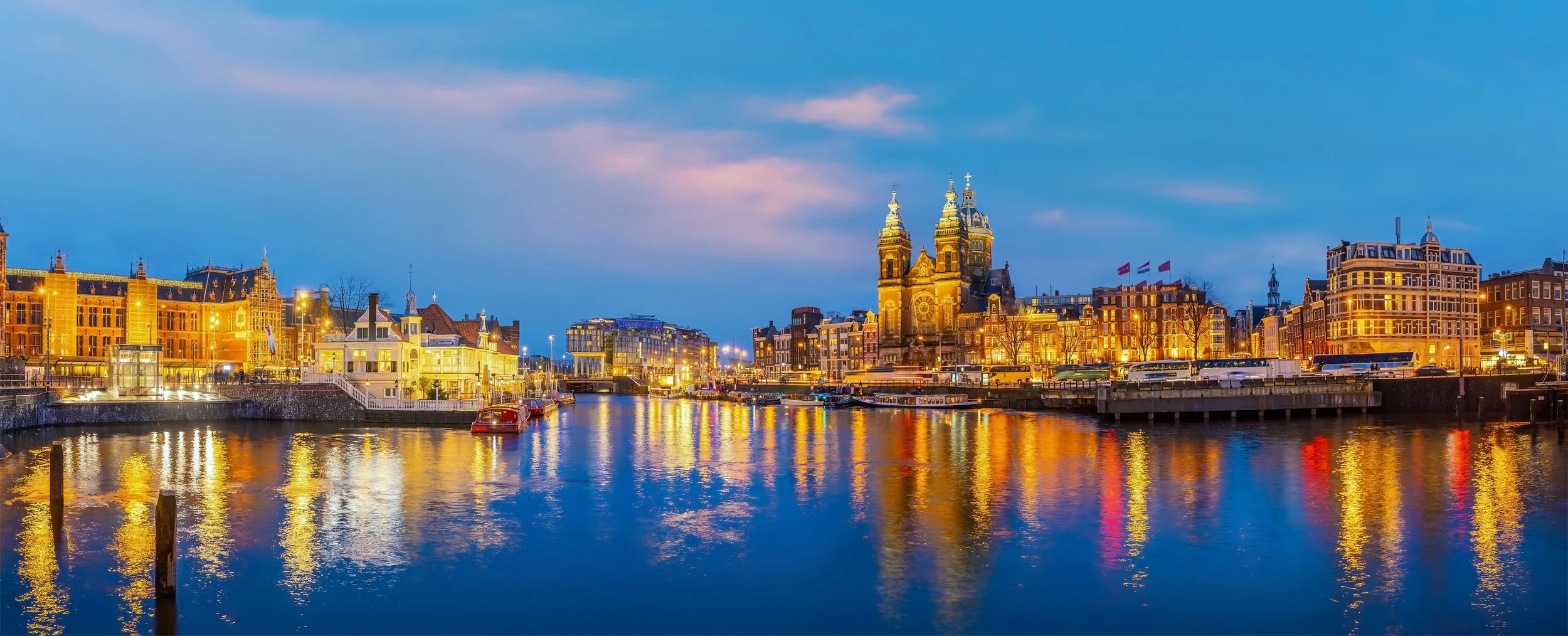 Nighttime view of Amsterdam cityscape with illuminated historic buildings and a church along a canal reflecting colorful lights on the water.