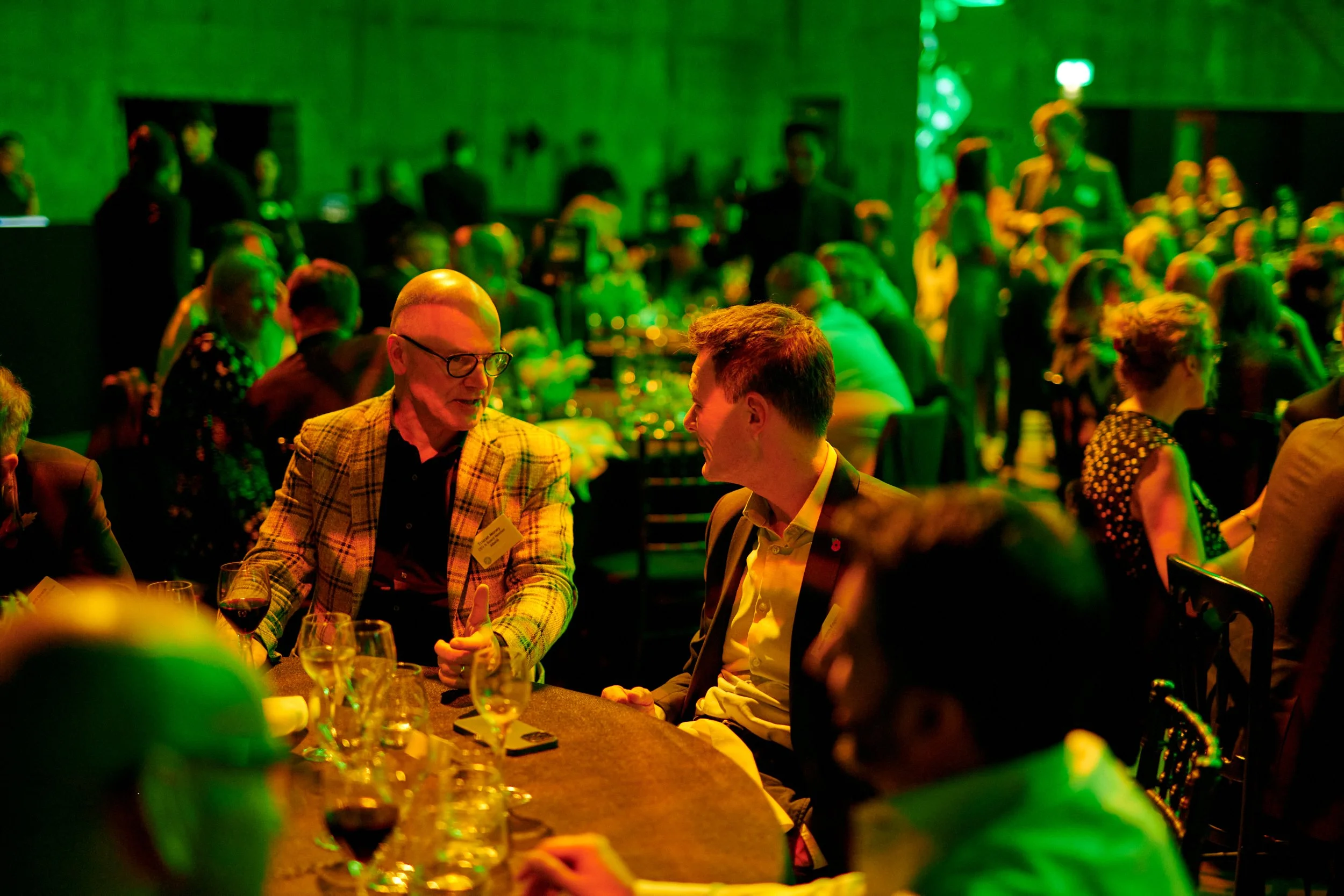 People socializing at a banquet or gala event in a dimly lit room with green lighting, with two men in conversation at a table with glasses of wine.