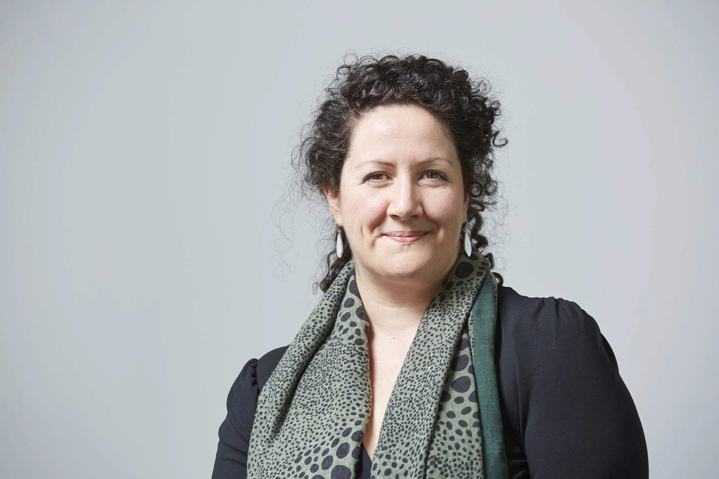 A woman with short curly dark hair, wearing a black top, a patterned scarf, and earrings, smiling against a light gray background.