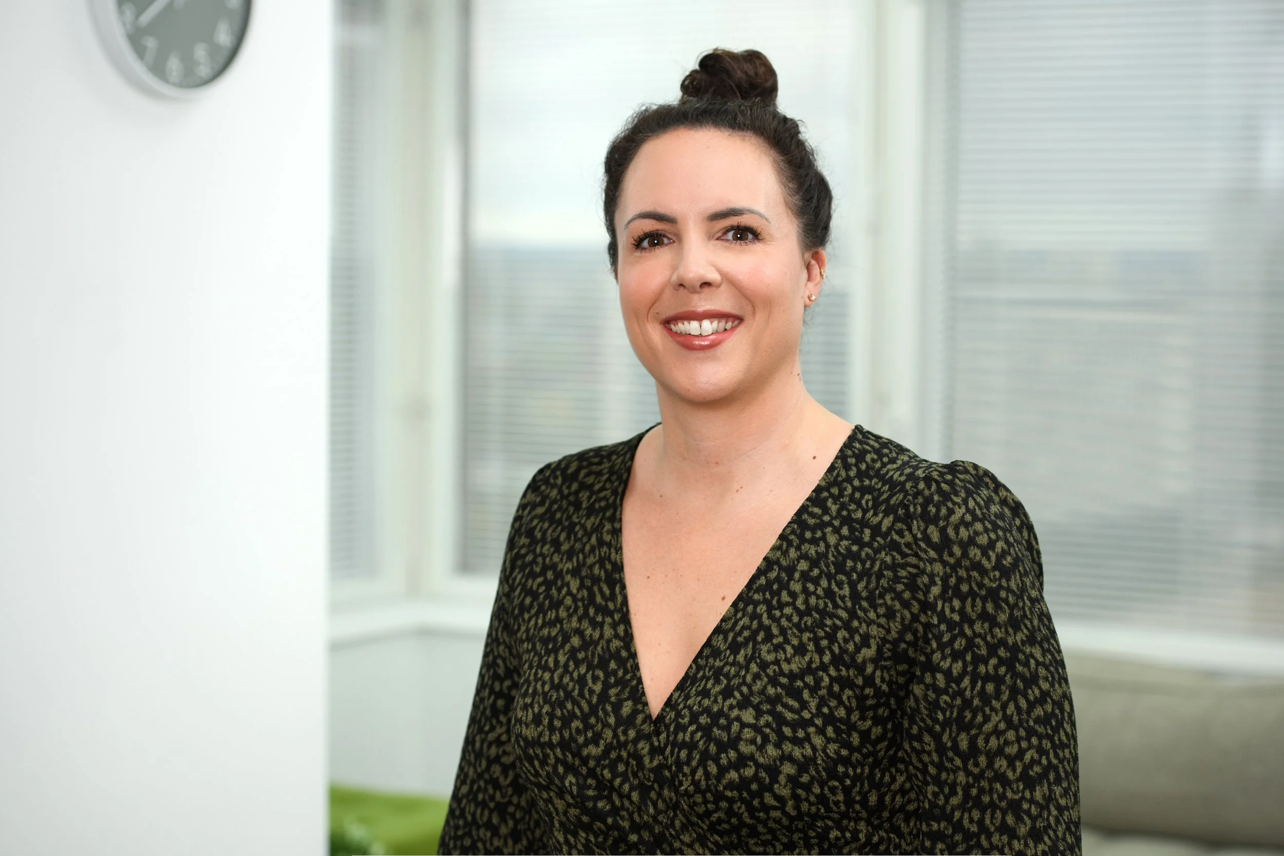 A woman with dark hair tied in a bun, smiling, wearing a low-cut patterned top, standing in a bright room with large windows and blinds.