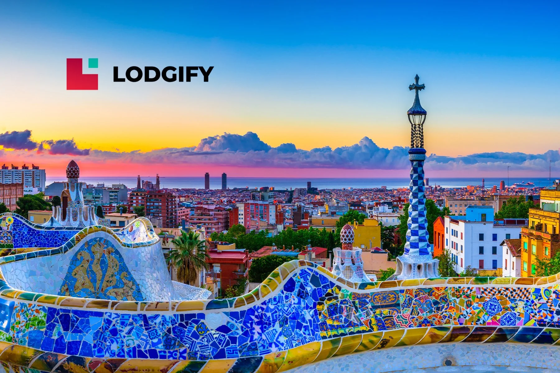 View of Barcelona at sunset, showcasing colorful mosaic benches and spires from Park Güell with cityscape and the sea in the background.
