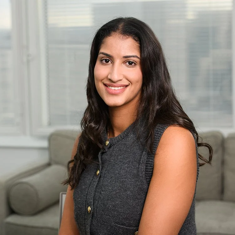 A young woman with long dark hair smiling, sitting in a modern office with large windows and a gray sofa in the background.