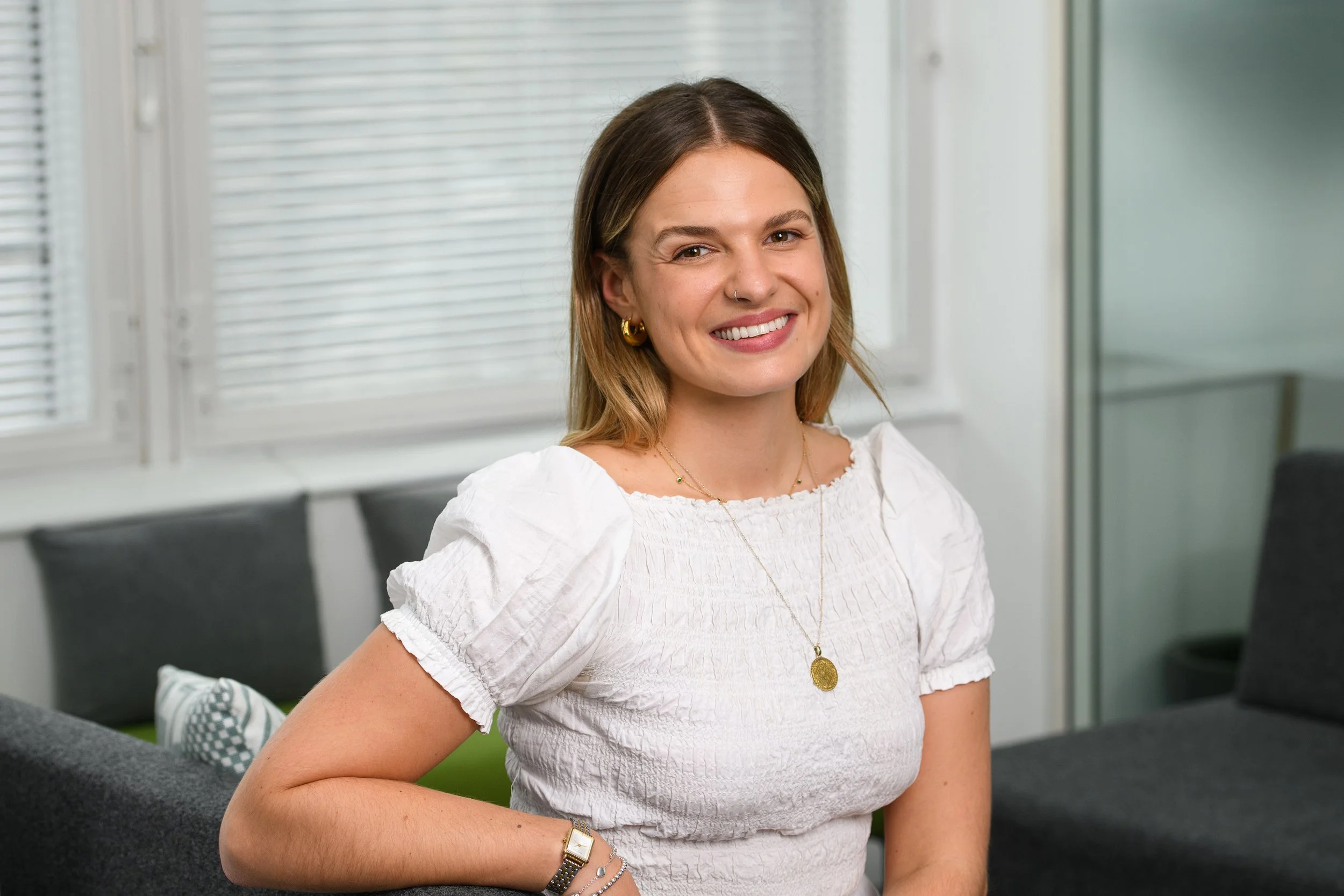 Young woman with medium-length brown hair smiling, wearing a white textured top, gold jewelry, and standing in a modern office with white blinds and a gray sofa.