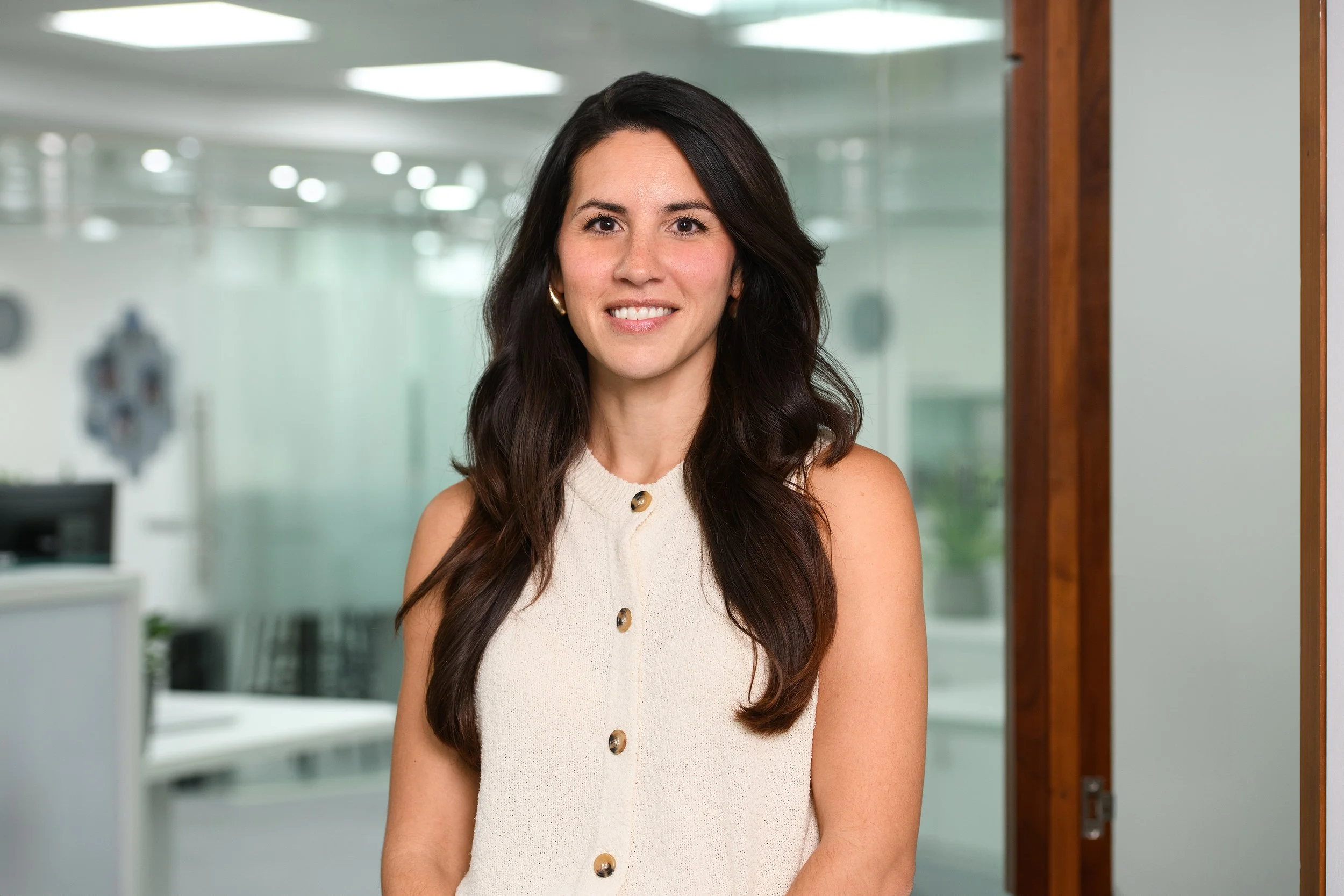 A woman with long dark hair, wearing a cream-colored sleeveless top with buttons, smiling in an office.