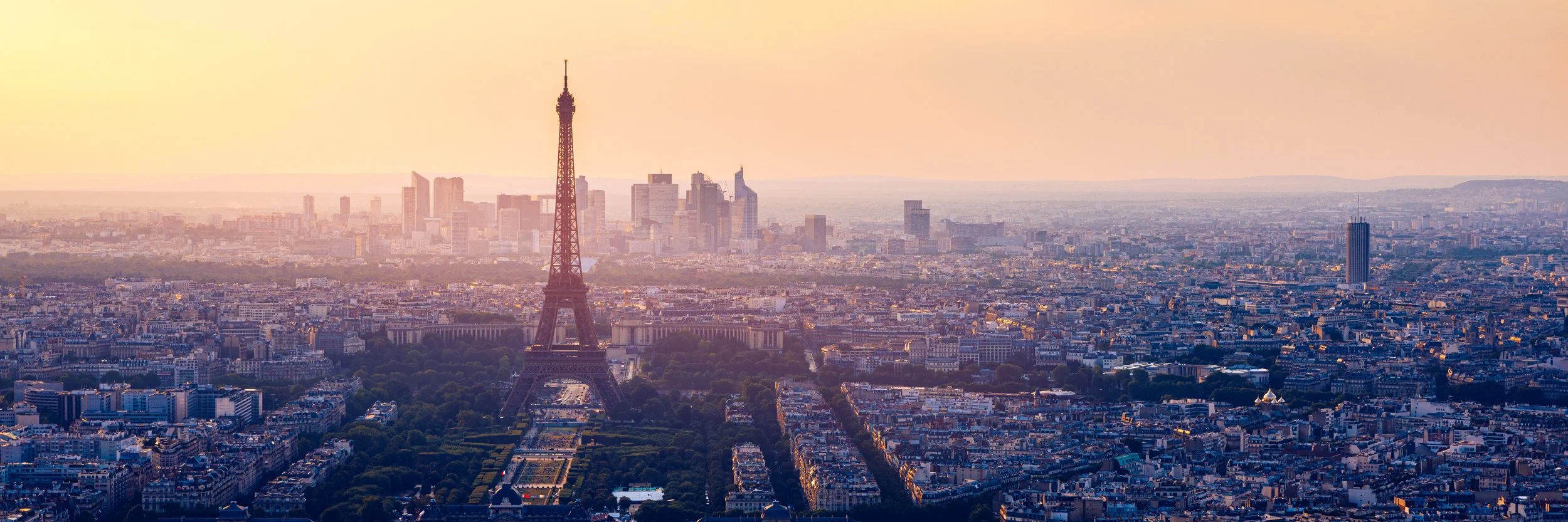 A panoramic view of Paris at sunset, featuring the Eiffel Tower in the foreground and the modern city skyline in the background.