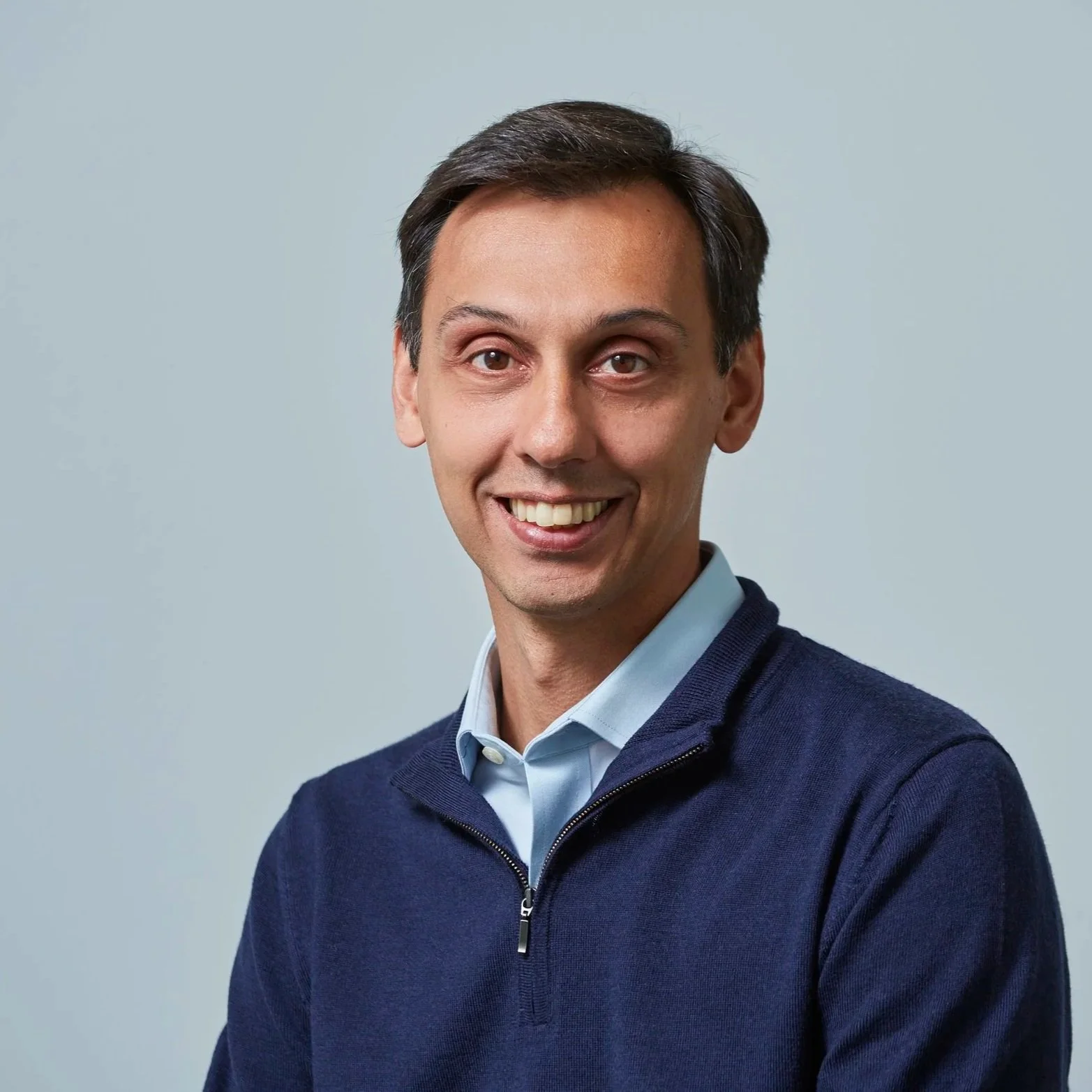 A smiling man with dark hair wearing a light blue shirt and navy blue sweater against a plain light gray background.
