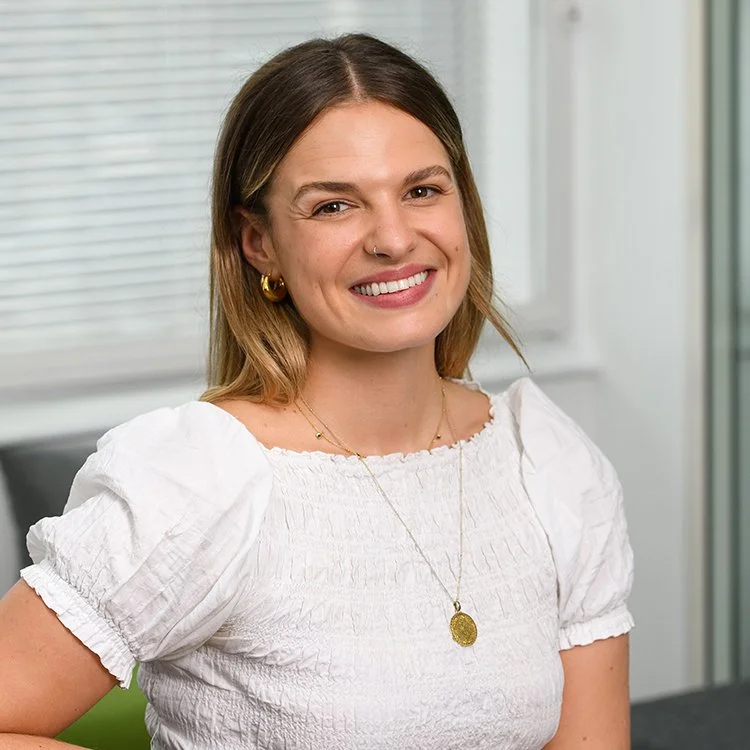 A woman with shoulder-length brown hair, wearing a white blouse, gold hoop earrings, a gold necklace, and a nose ring, smiling in an indoor setting with a window and a green chair in the background.