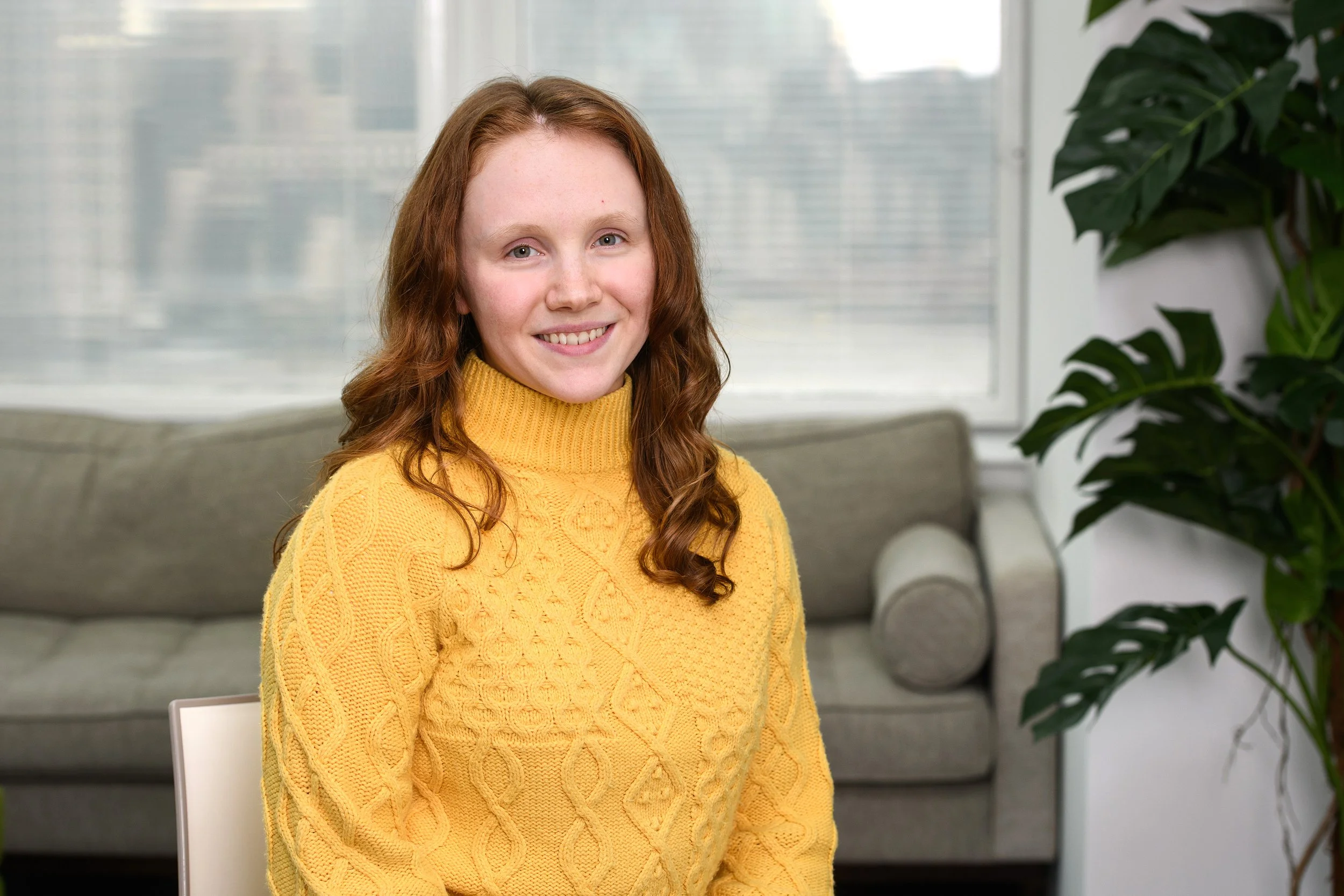 A young woman with long, wavy red hair and fair skin, wearing a yellow cable-knit turtleneck sweater, smiling and sitting in a room with large windows, a gray sofa, and a large green potted plant.