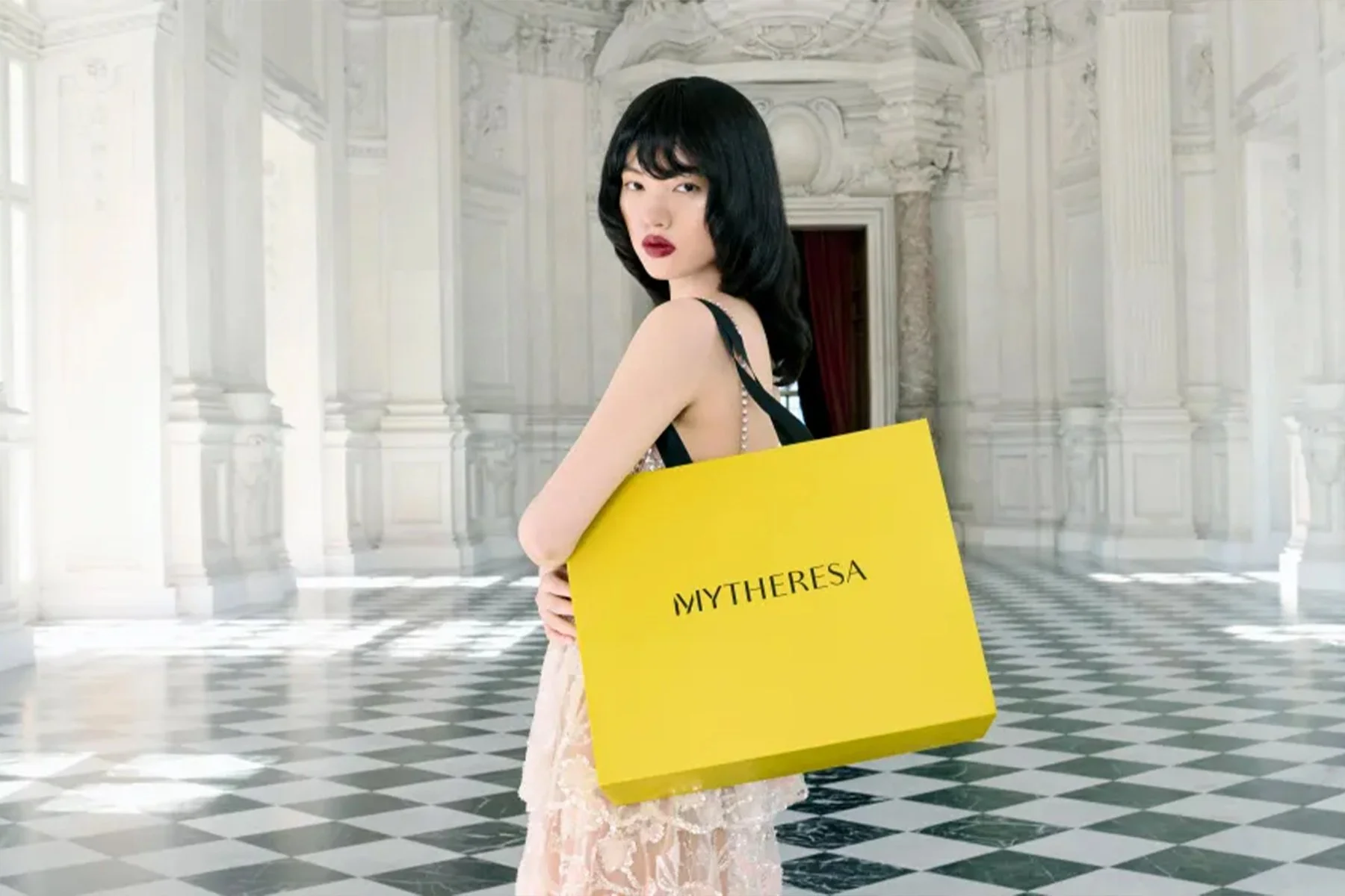 A woman with dark hair in a black dress holding a large yellow shopping bag with the word 'MYTHERESA' written on it, standing in a grand, white marble hall with checkered black and white floor and ornate architectural details.