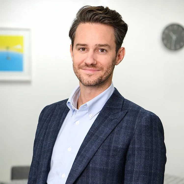A man with dark brown hair and a beard, smiling in an office setting, wearing a dark suit jacket and light blue collared shirt, blurred clock and artwork in the background.