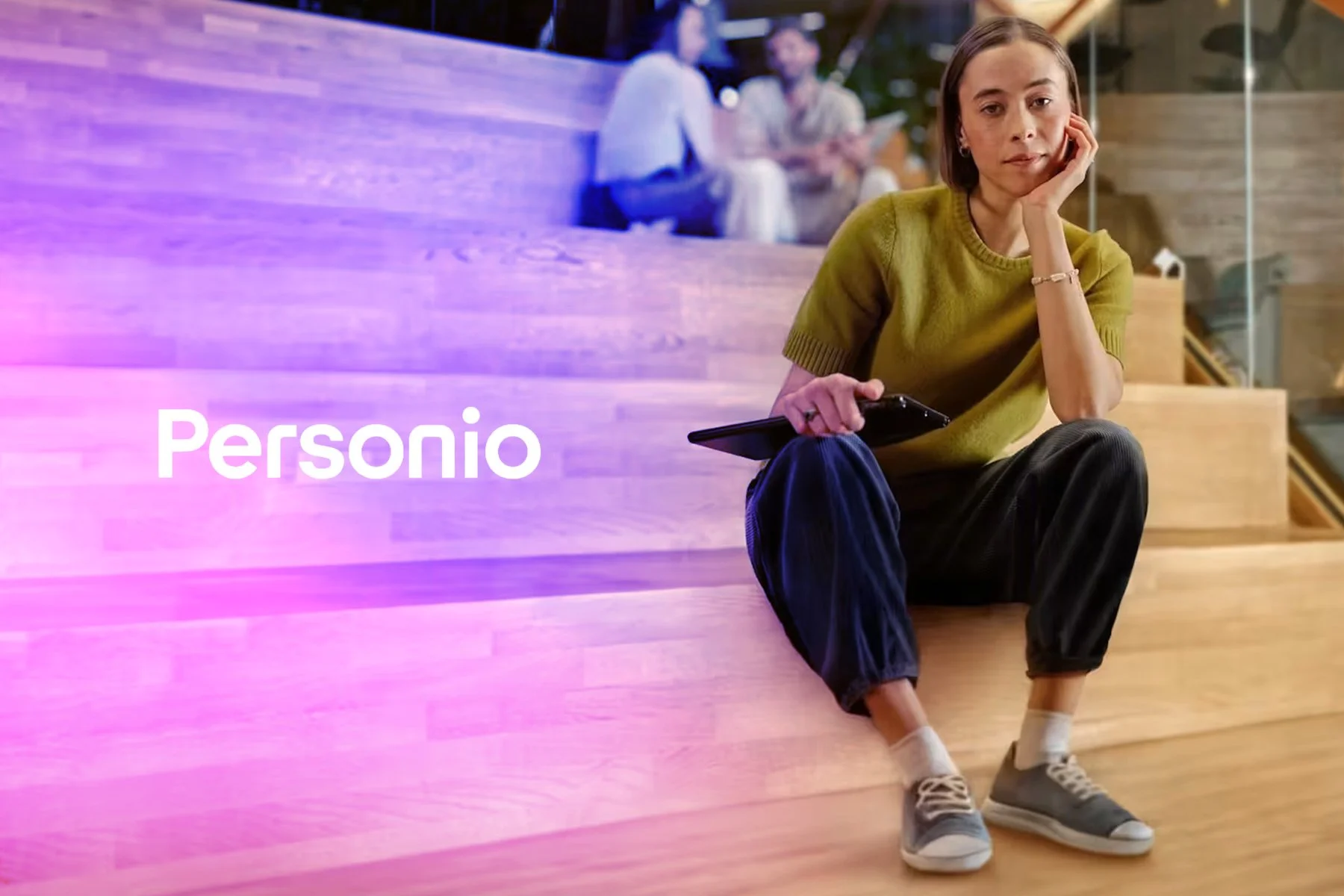 A young woman sitting on wooden stairs in a modern indoor space, holding a phone, wearing a yellow-green sweater, black pants, and sneakers, with her head resting on her hand, and two people blurred in the background. The word 'Personio' is visible on the left side of the image.