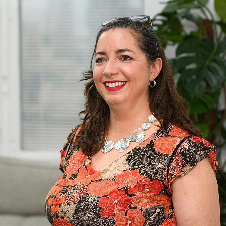 A woman with medium-length brown hair wearing a floral dress with orange, beige, and black patterns, smiling, with a large silver necklace and earrings, standing in front of a window and a plant.
