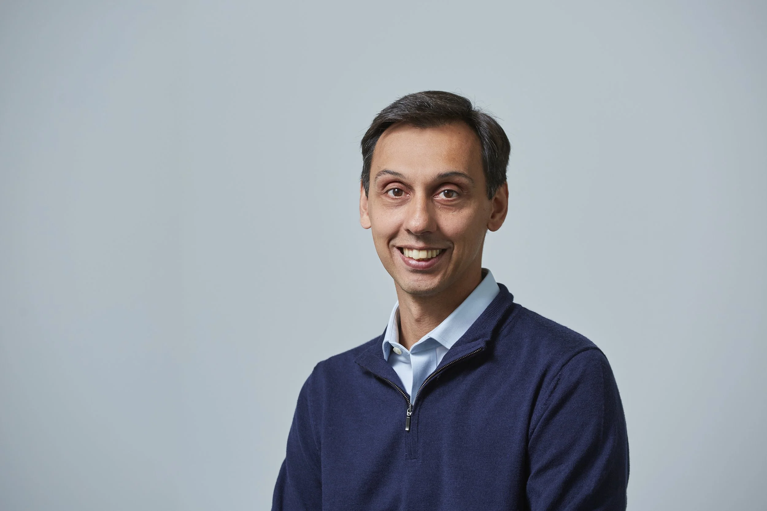 A man with dark hair, wearing a blue sweater over a light blue collared shirt, smiling at the camera against a light gray background.