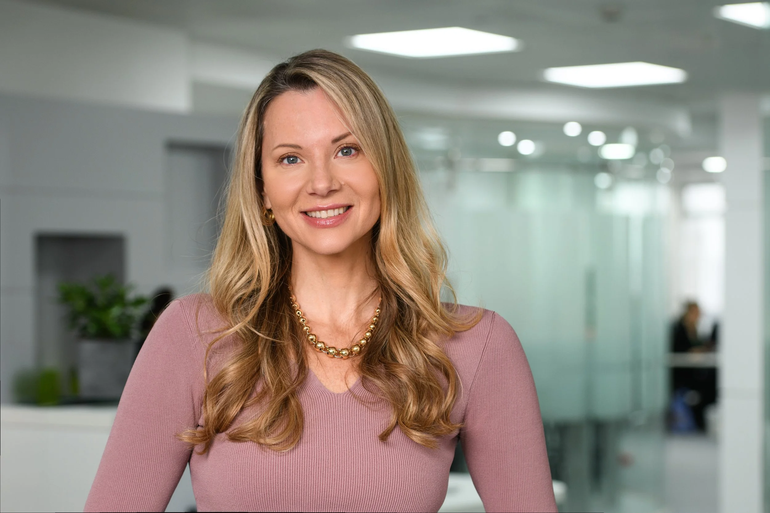 A woman smiling in an office hallway with modern lighting and potted plants in the background.