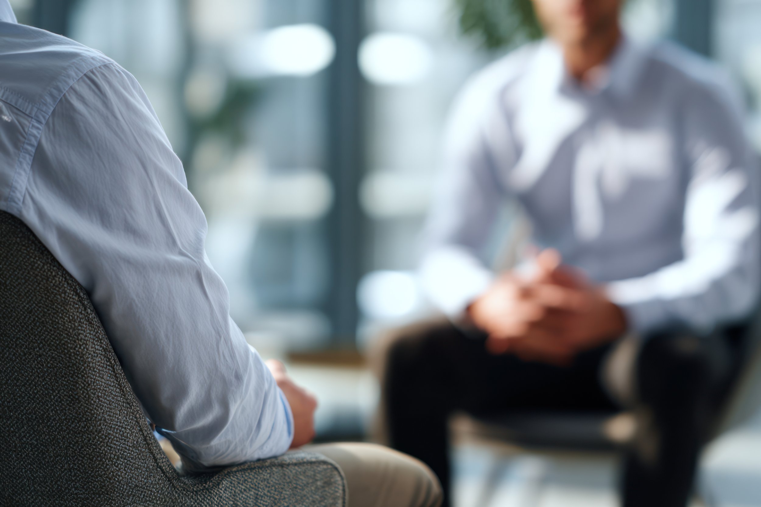 Close-up of a person wearing a white shirt sitting in a waiting area, with another person blurred in the background, possibly using a phone or device. The setting appears to be a modern office or medical facility.